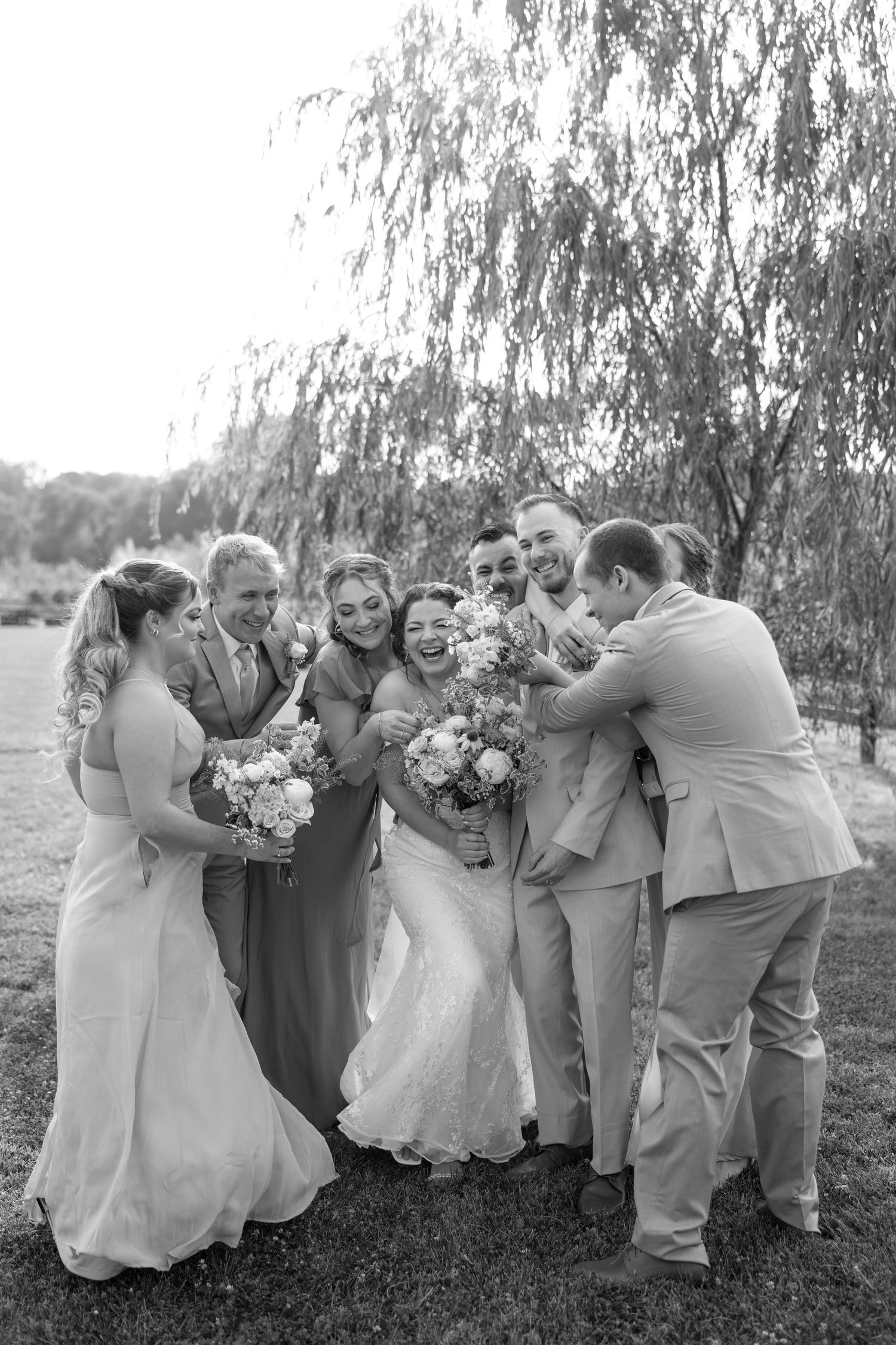 Wedding party celebrates joyfully under weeping willow trees during outdoor reception at Kin Lock wedding.