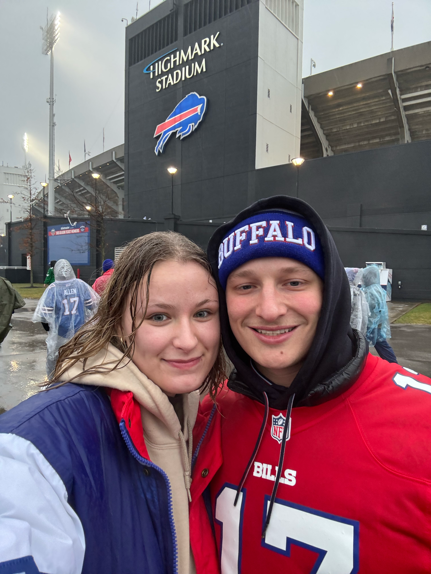 Two Buffalo Bills fans smiling in front of Highmark Stadium on a rainy game day.