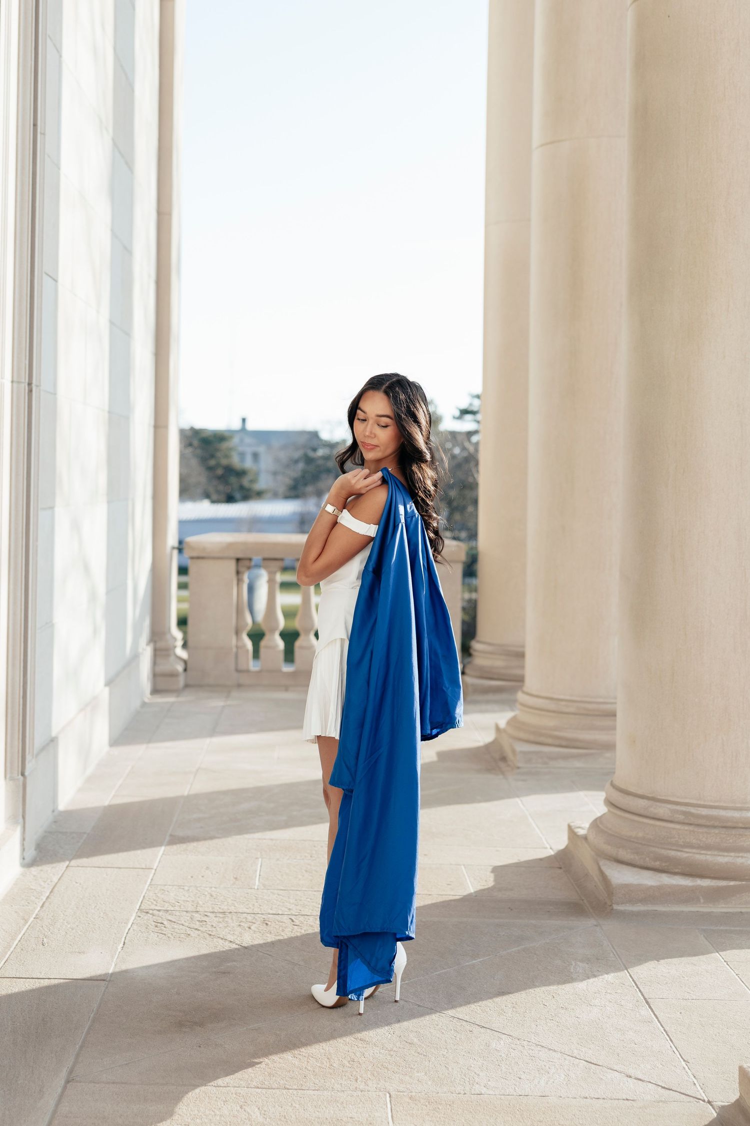Portrait series featuring someone in a white dress holding their UB blue graduation gown posing between white classical columns.