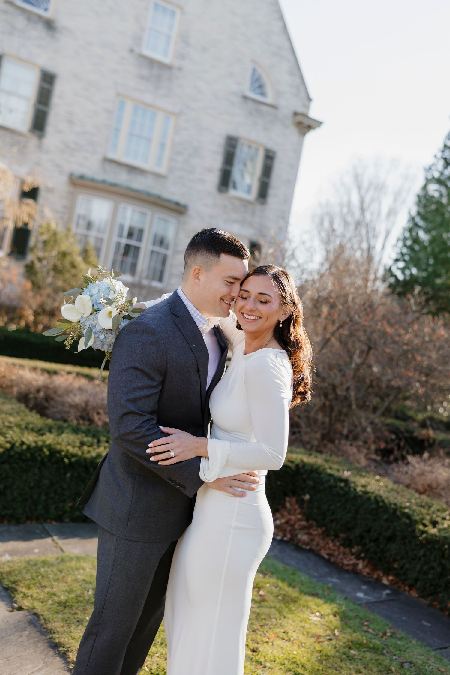 A couple embracing outside a white stone house during a winter wedding photoshoot at George eastman house for Rochester NY elopement.
