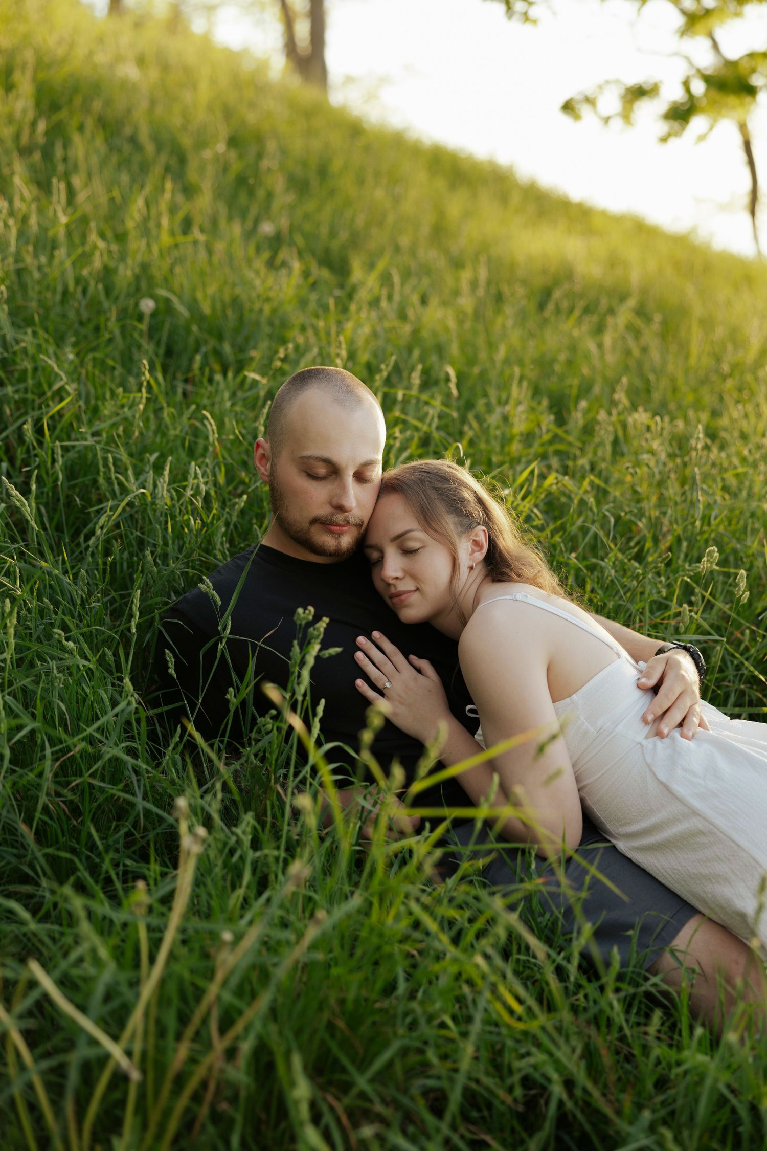 A couple shares an intimate moment while lying in tall grass on a sunlit hillside during golden hour.