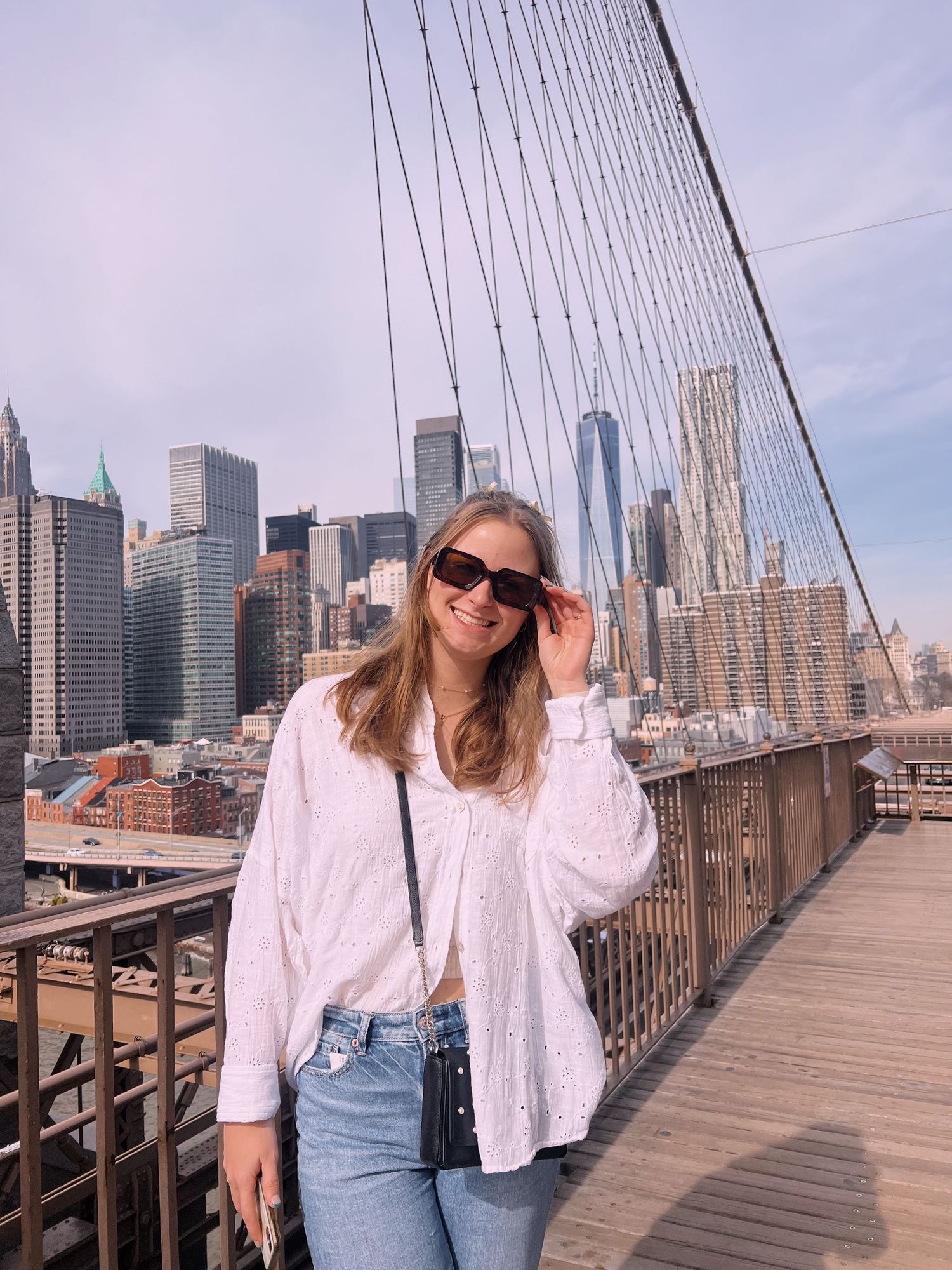 Woman in white shirt and jeans posing on Brooklyn Bridge with NYC skyline in background on sunny day.