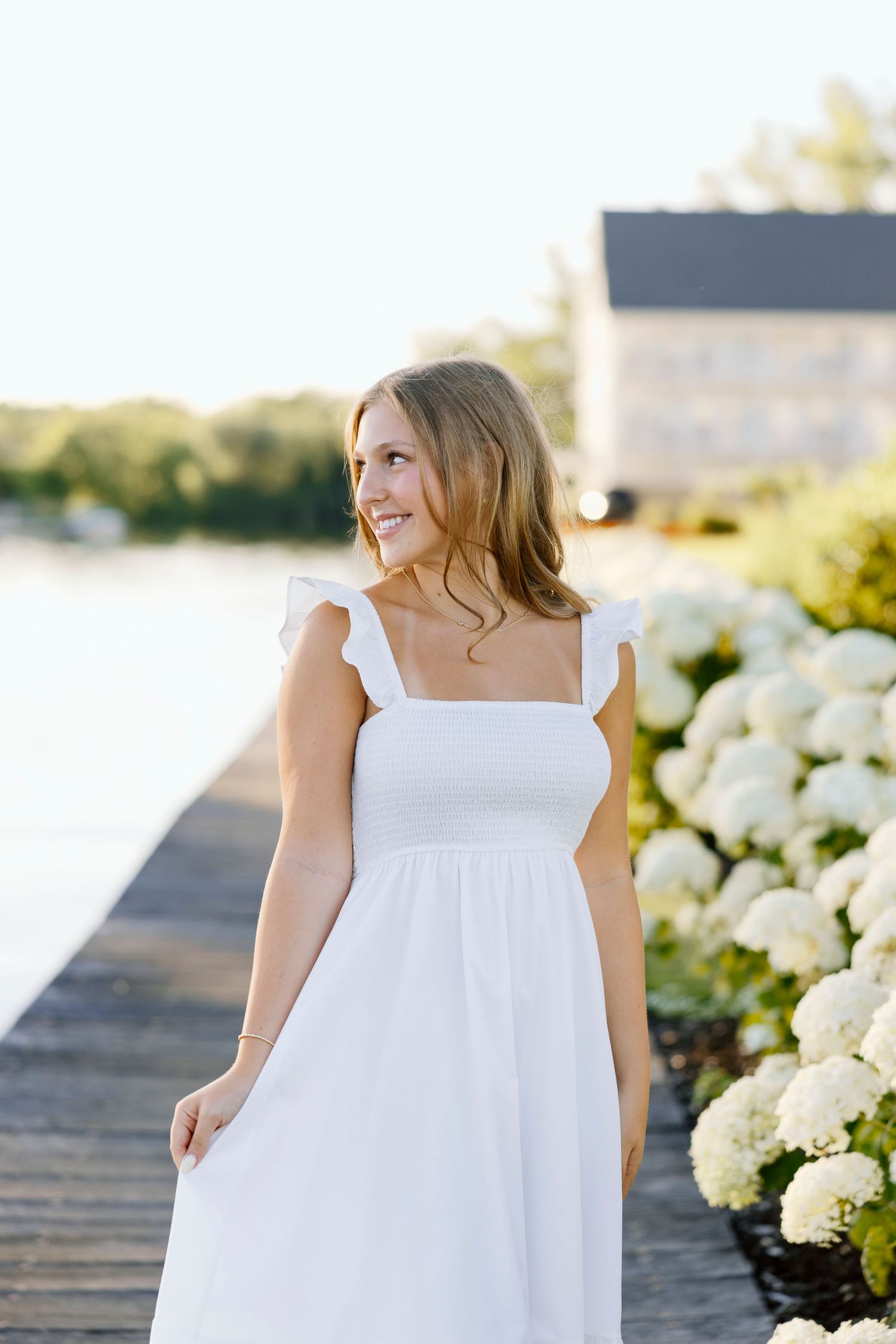 High School senior girl in white dress stands near blooming hydrangea bushes at sunset during senior portaits in Rochester, NY.