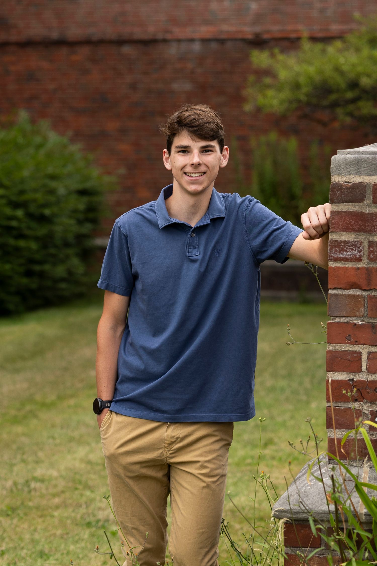 High school senior boy in navy polo and khaki pants poses by brick column outdoor during senior portraits in Rochester, NY.