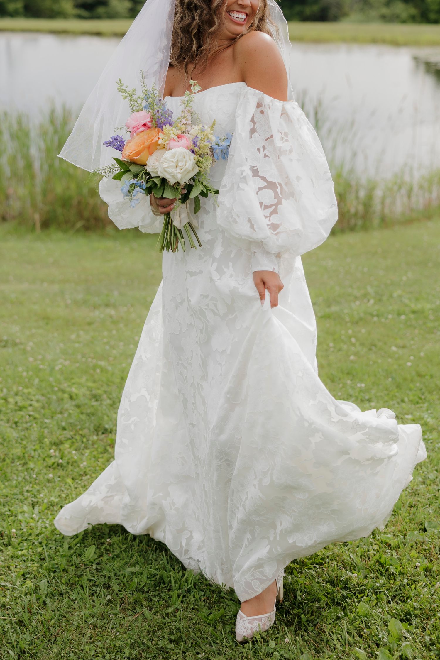 Bride in off-shoulder lace gown with puffy sleeves holds colorful spring bouquet by lakeside at Pennsylvania barn wedding.
