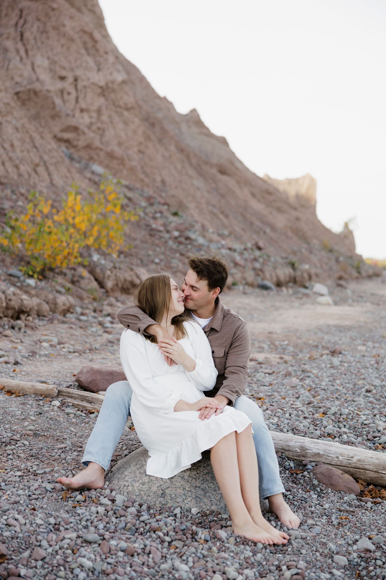 Couple shares intimate moment on rocky beach against dramatic cliff backdrop at Chimney Bluffs State Park engagement photos.