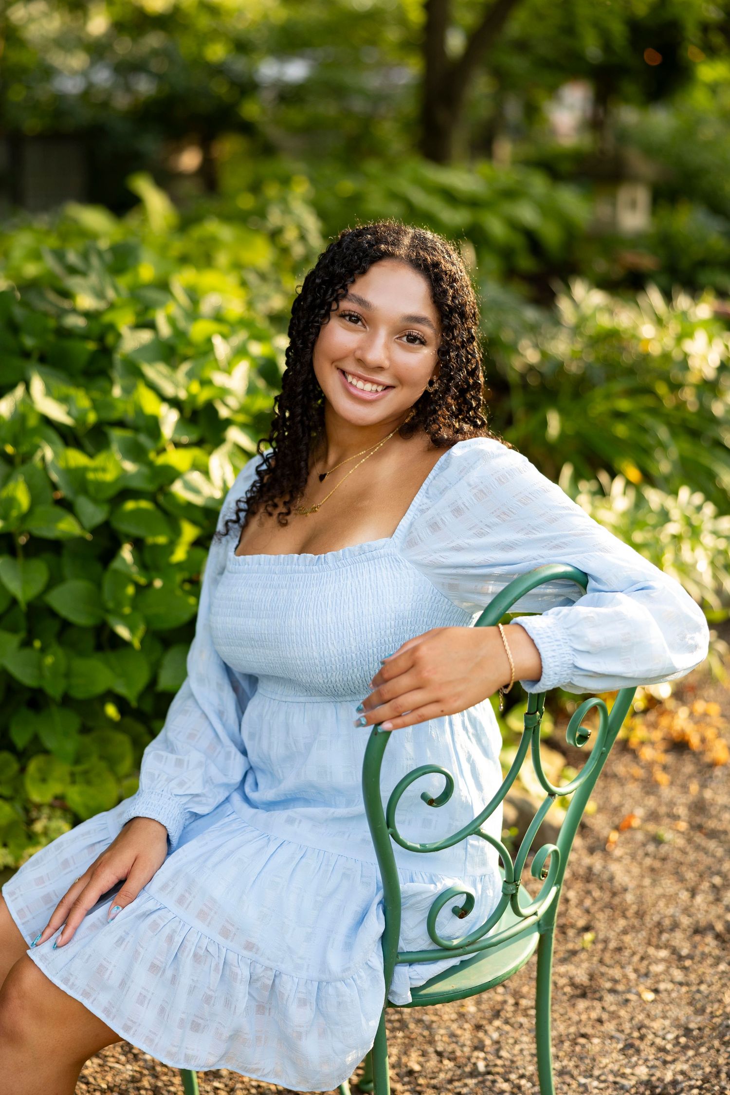 High School senior girl in light blue ruffled top poses on vintage garden chair outdoors at Sara's Garden for senior portraits, Rochester NY