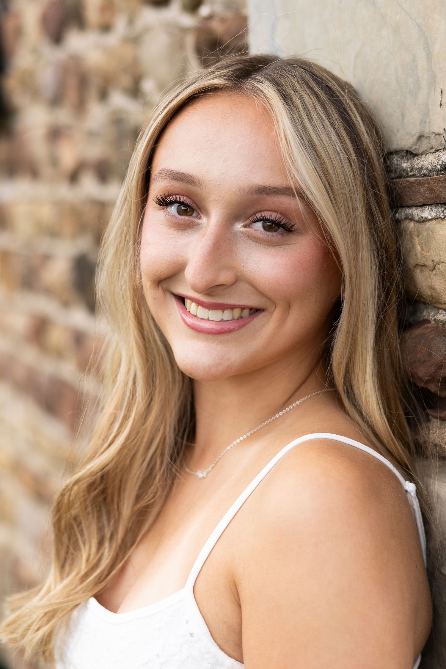 Blonde High school senior portaits against a cobblestone wall in a white dress at Tinker Nature Center, Rochester, NY.