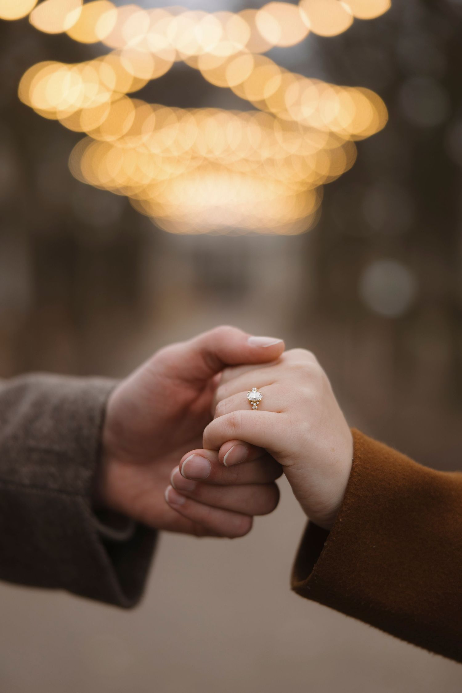 Two hands clasped together with an engagement ring sparkling under warm bokeh lights at Memorial Art Gallery, Rochester engagement photos.