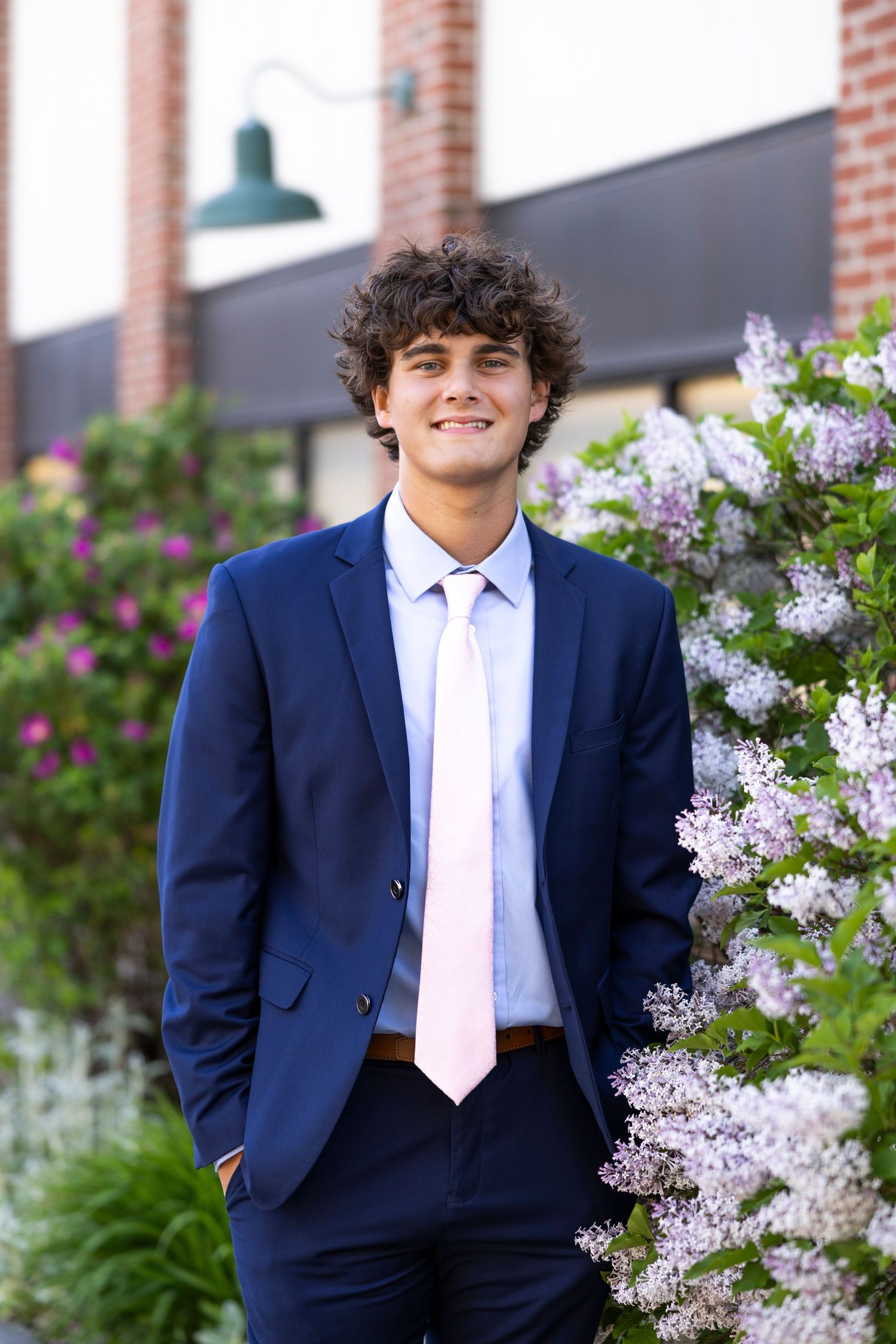 A formal portrait against flowering lilac bushes wearing a navy suit and pink tie at High Falls for Rochester NY senior portaits.
