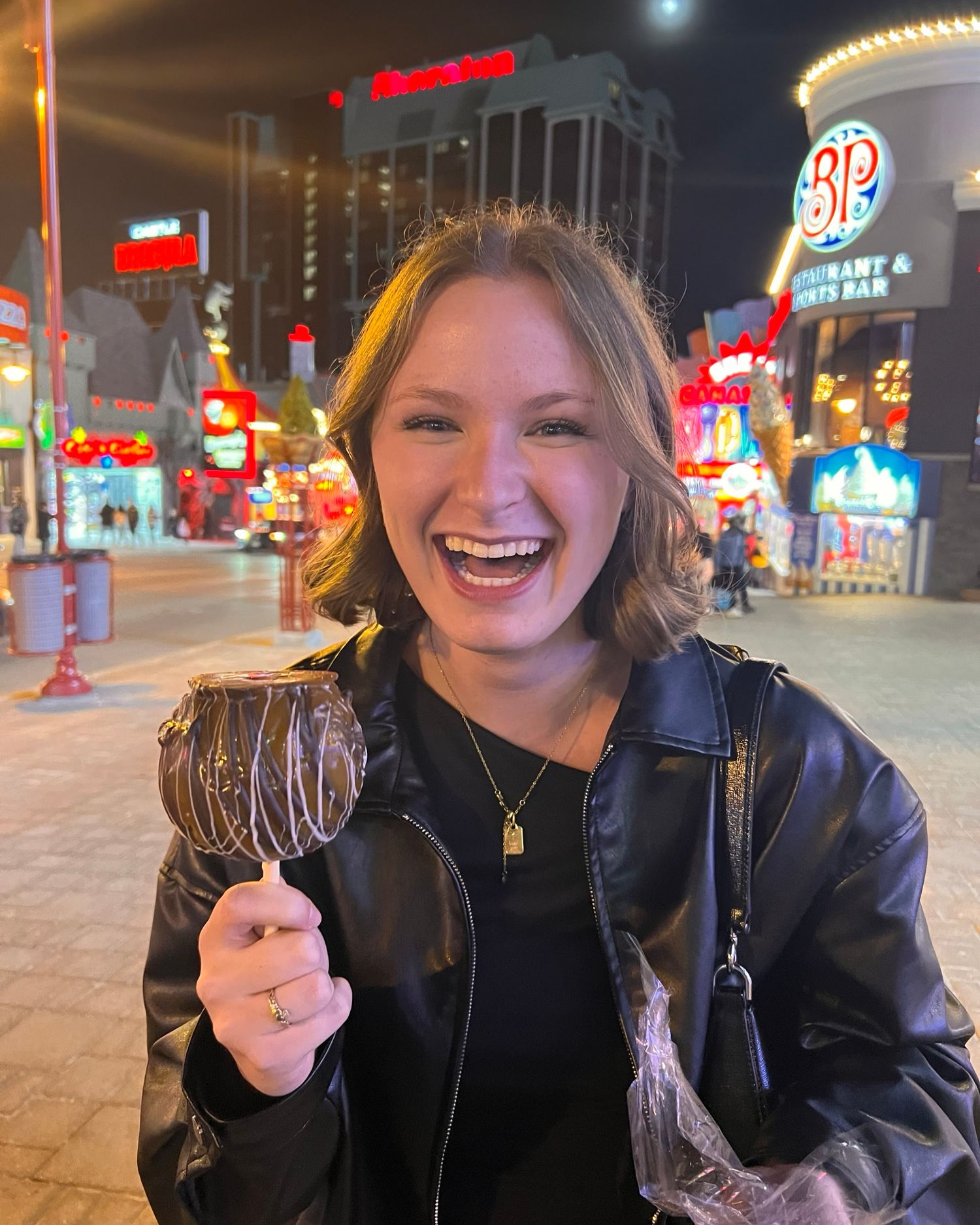 Smiling young woman holding a chocolate-dipped candy apple on a vibrant city street at night.