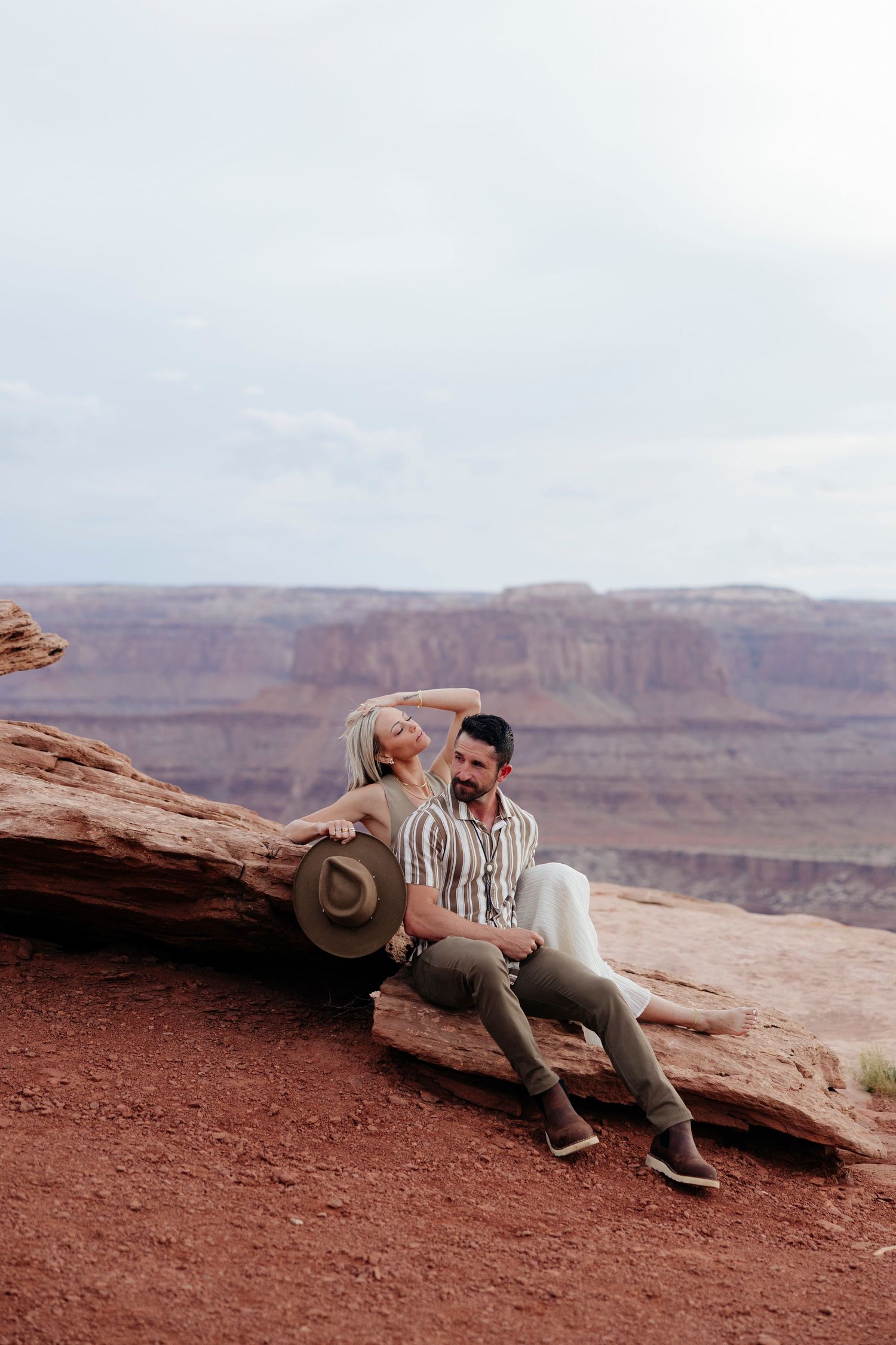 A couple sits together on red rocks overlooking the vast Grand Canyon landscape at sunset.