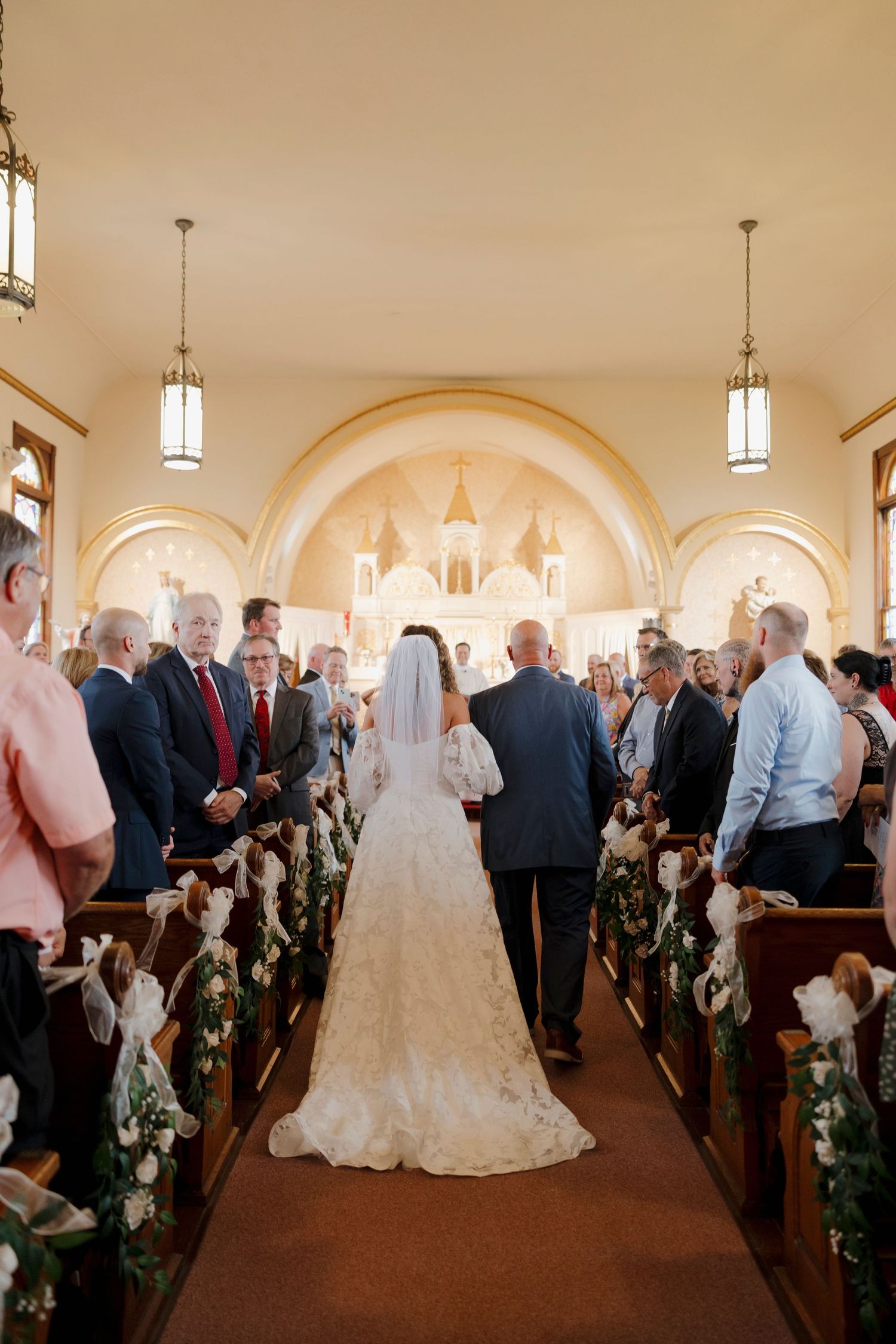 Wedding ceremony in a traditional church with arched ceilings and guests lining the aisle as bride and father walk down. Rochester, NY