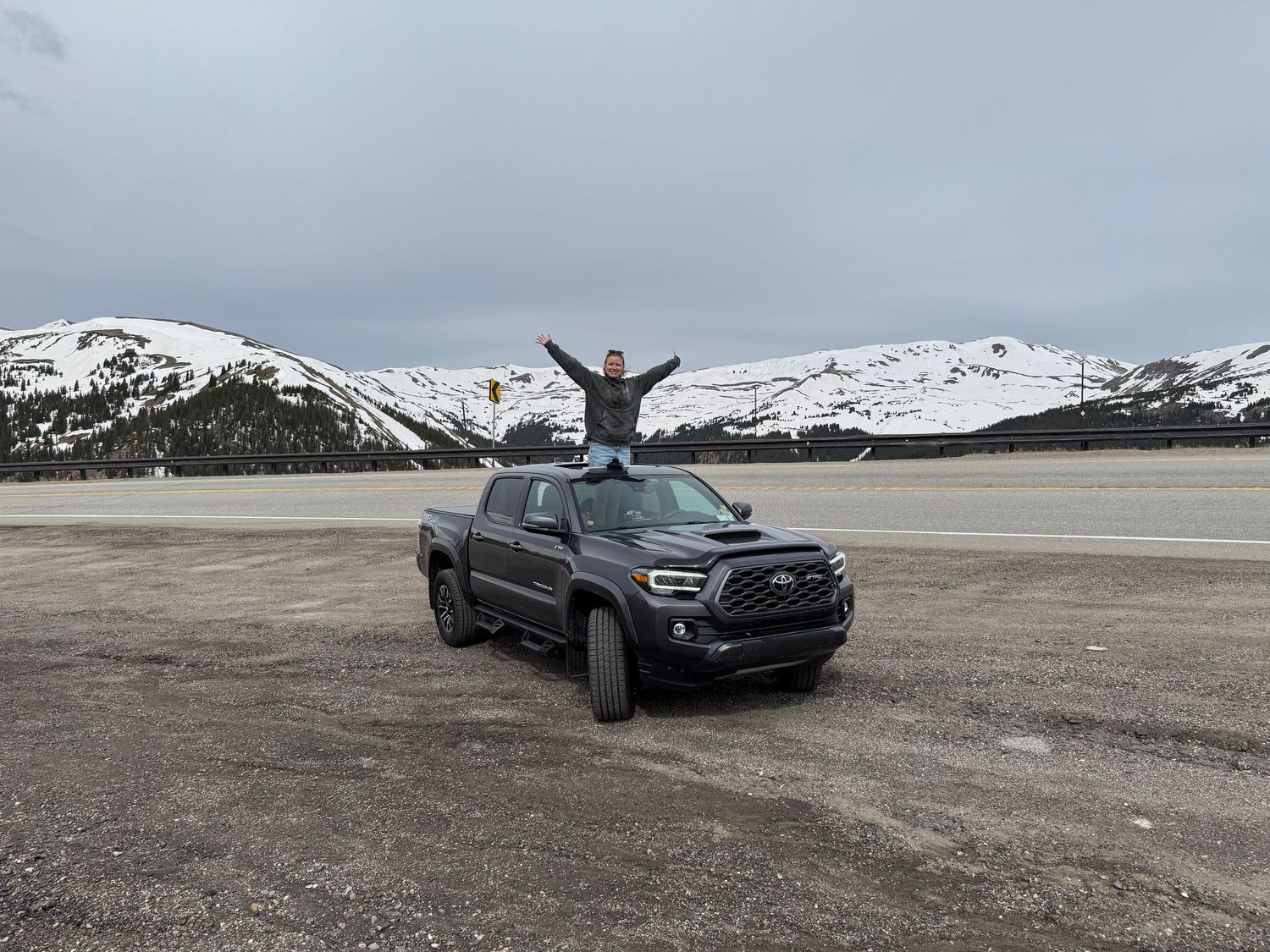 Person standing on top of a Toyota Tacoma truck in a snowy mountain landscape with arms raised.