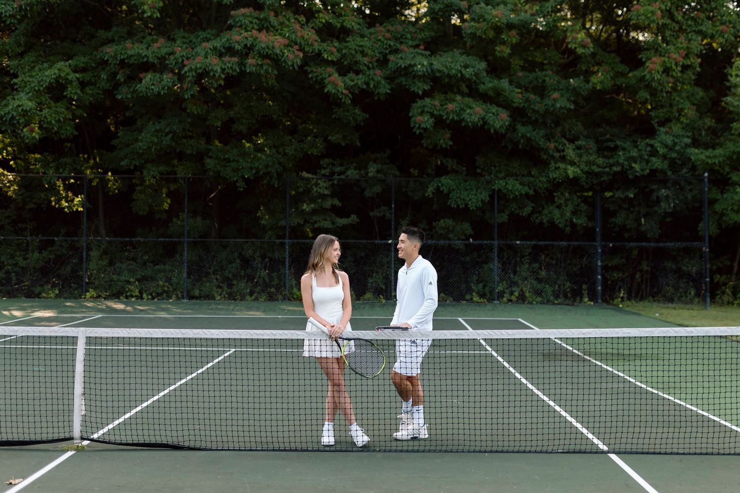 Two people in white tennis outfits standing on court having conversation for tennis themed engagement photos, Rochester, NY.
