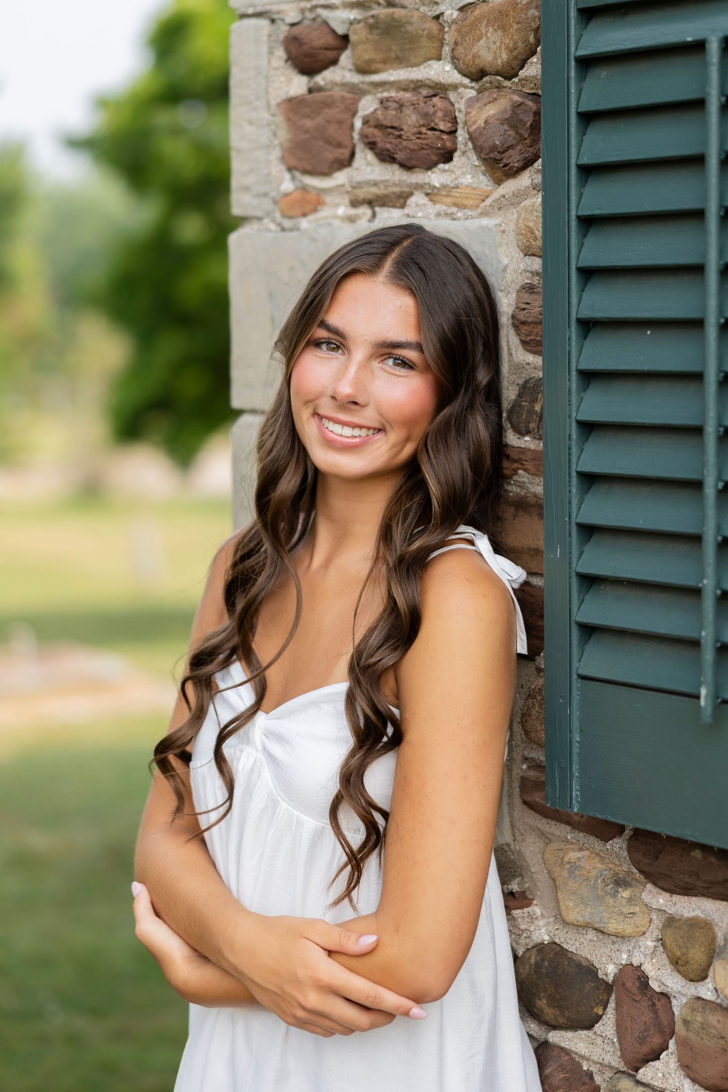 Casual senior portraits against stone wall with green shutters in natural lighting at Tinker Nature Center in Rochester, NY.