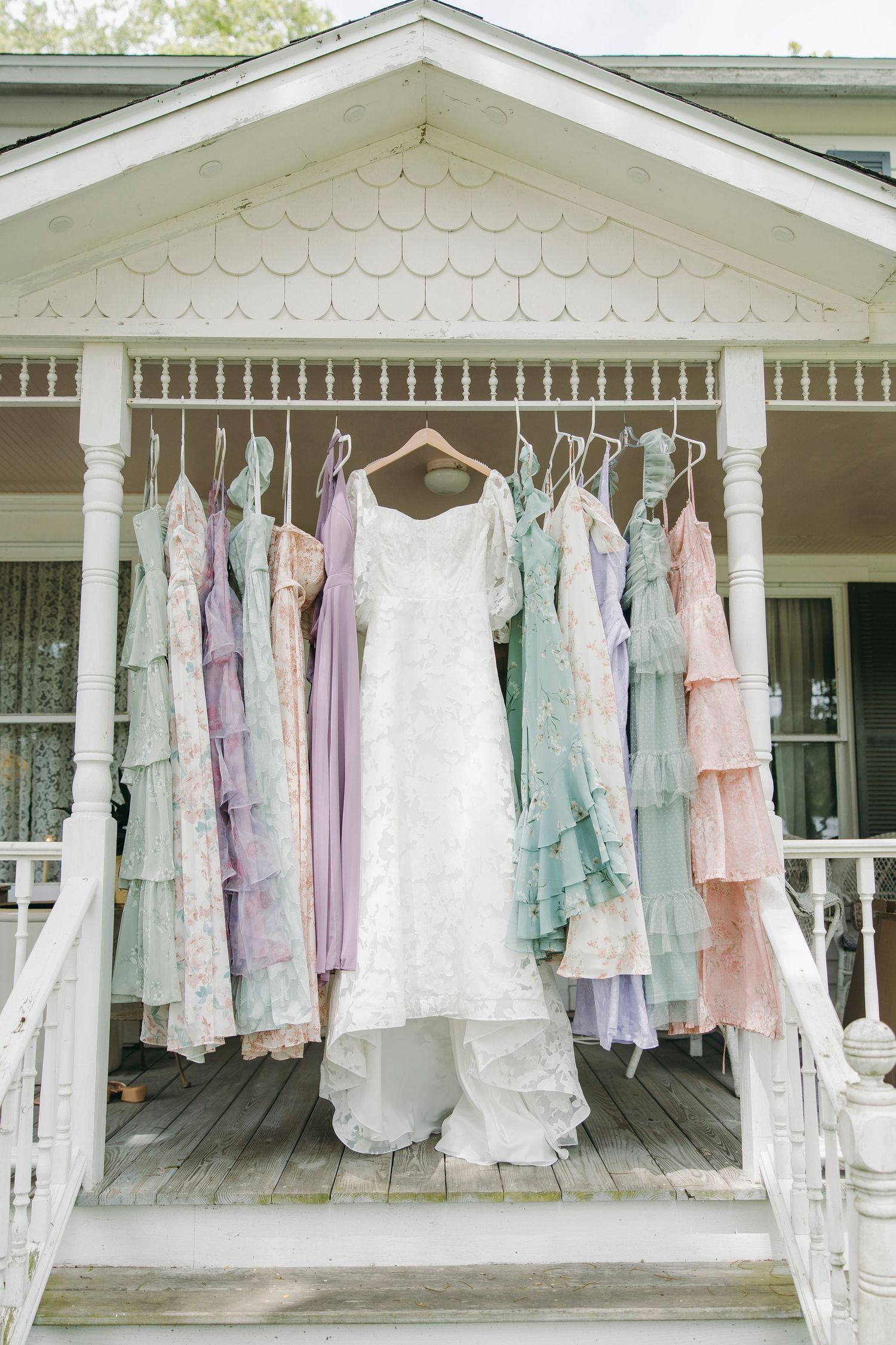 Pastel garden party themed bridesmaid dresses and white wedding gown hanging on farmhouse porch railing in natural light, Rochester, NY.