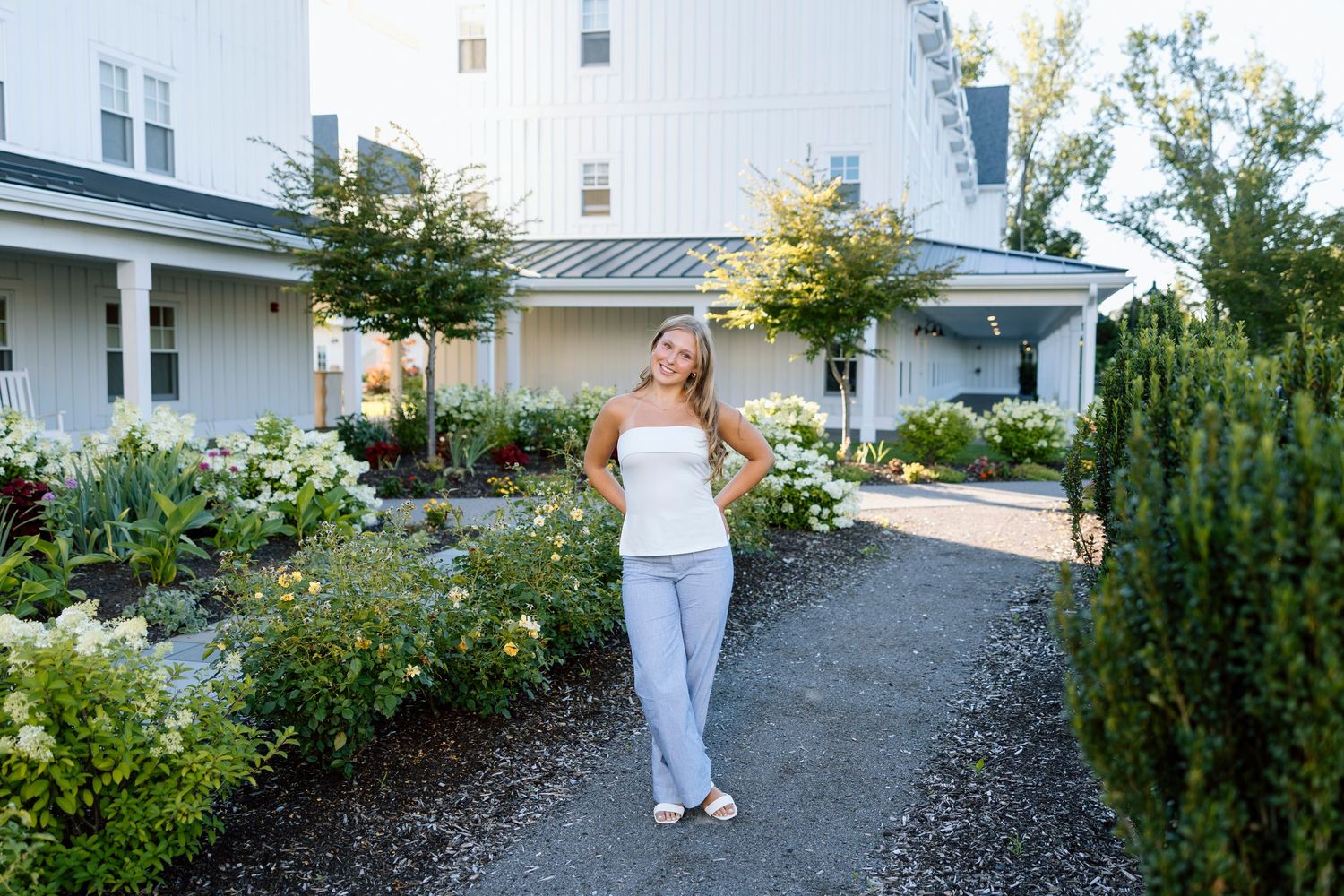 Senior portait standing in garden pathway with landscaped shrubs and white trim architecture in summer setting at Canadaigua Lake House.