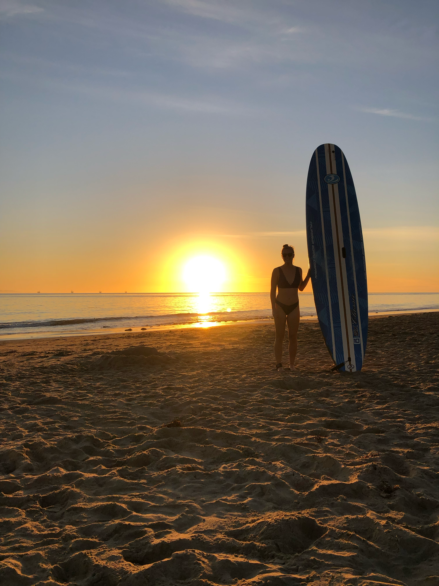 Silhouette of a surfer holding a longboard on a sandy beach during a golden sunset over the ocean.