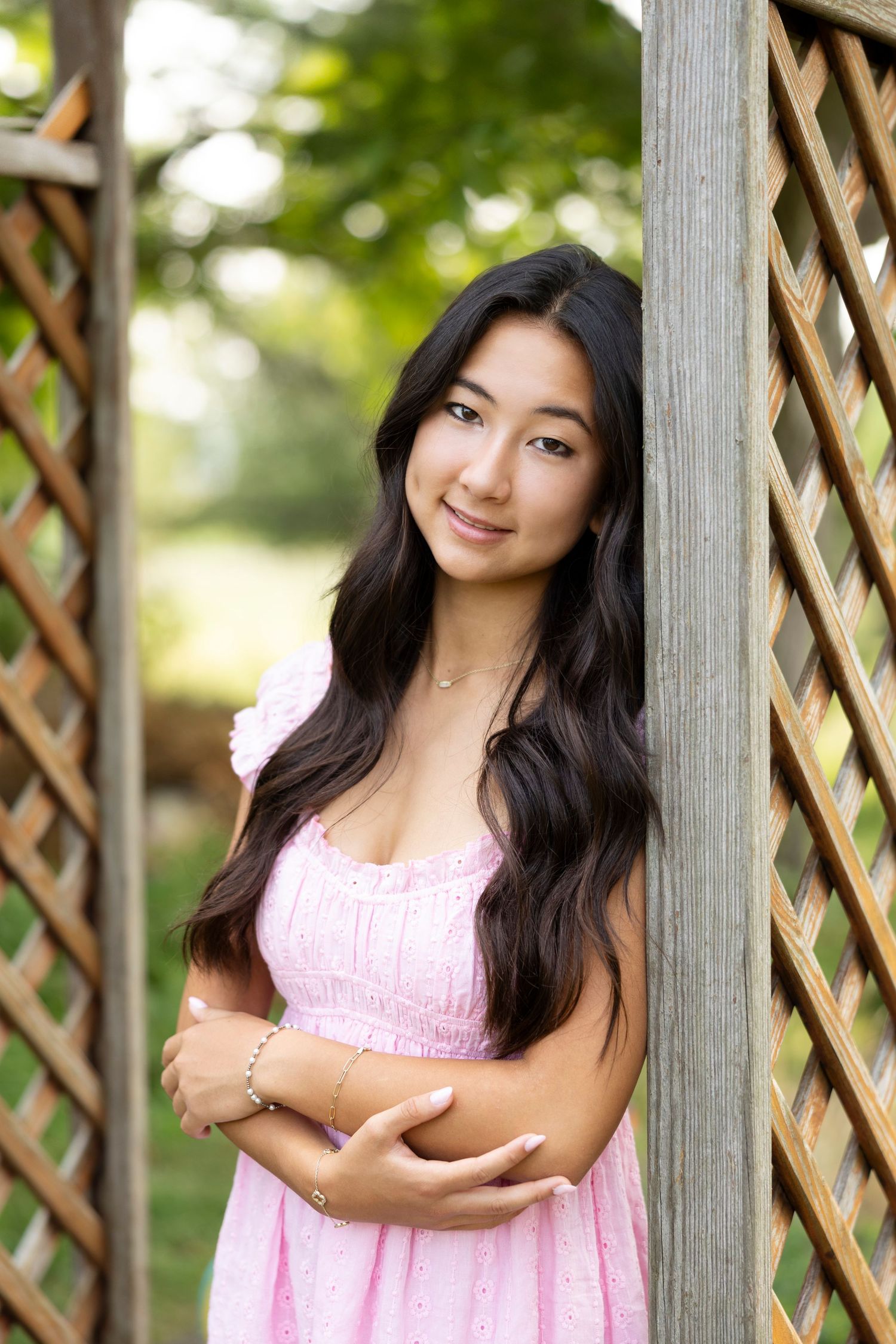 Natural light portrait against wooden lattice in pink smocked summer dress during senior photos at Tinker Nature Center. Rochester, NY.