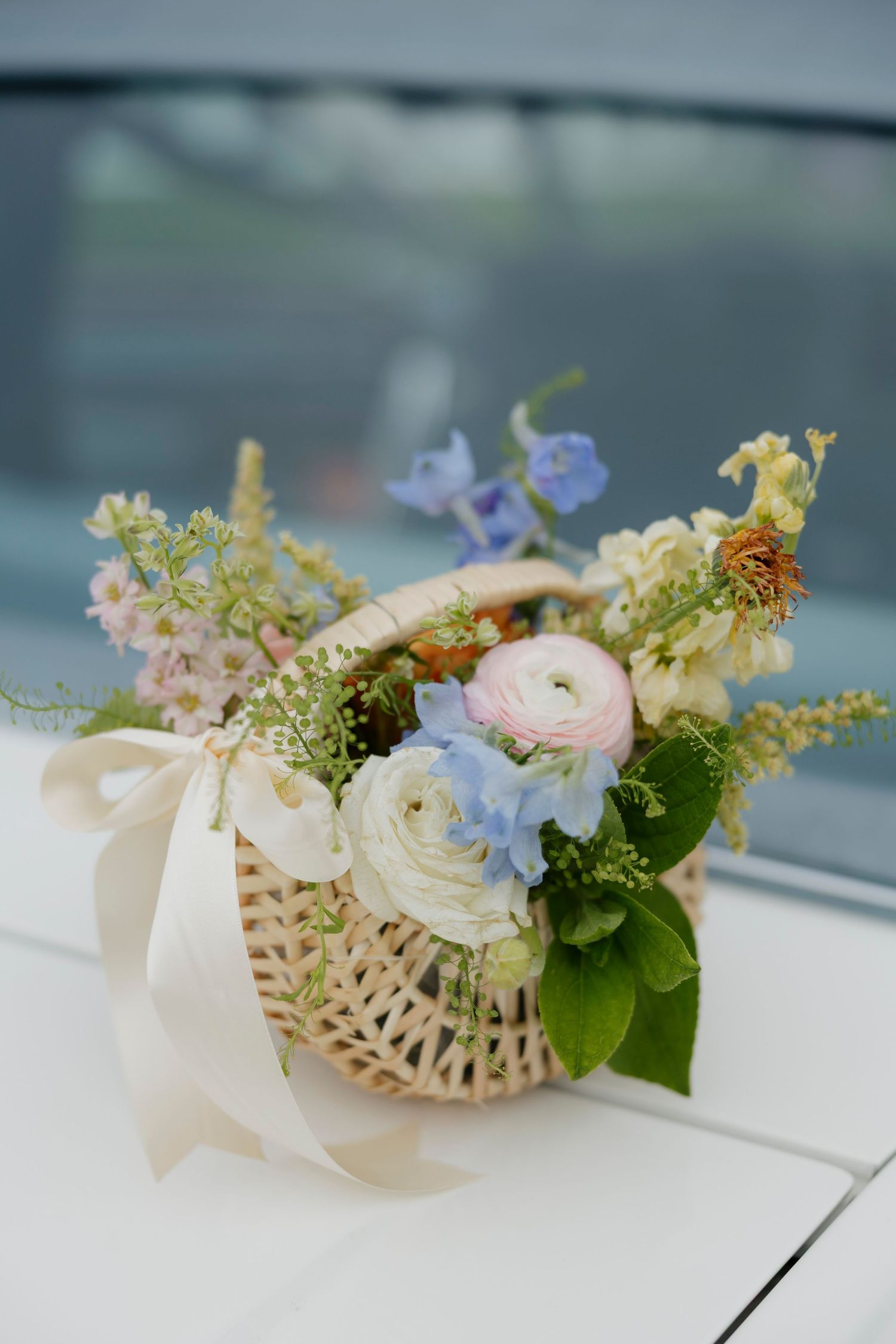 Delicate garden party floral arrangement with pastel ranunculus and blue flowers in woven basket with ribbon in Rochester, NY.