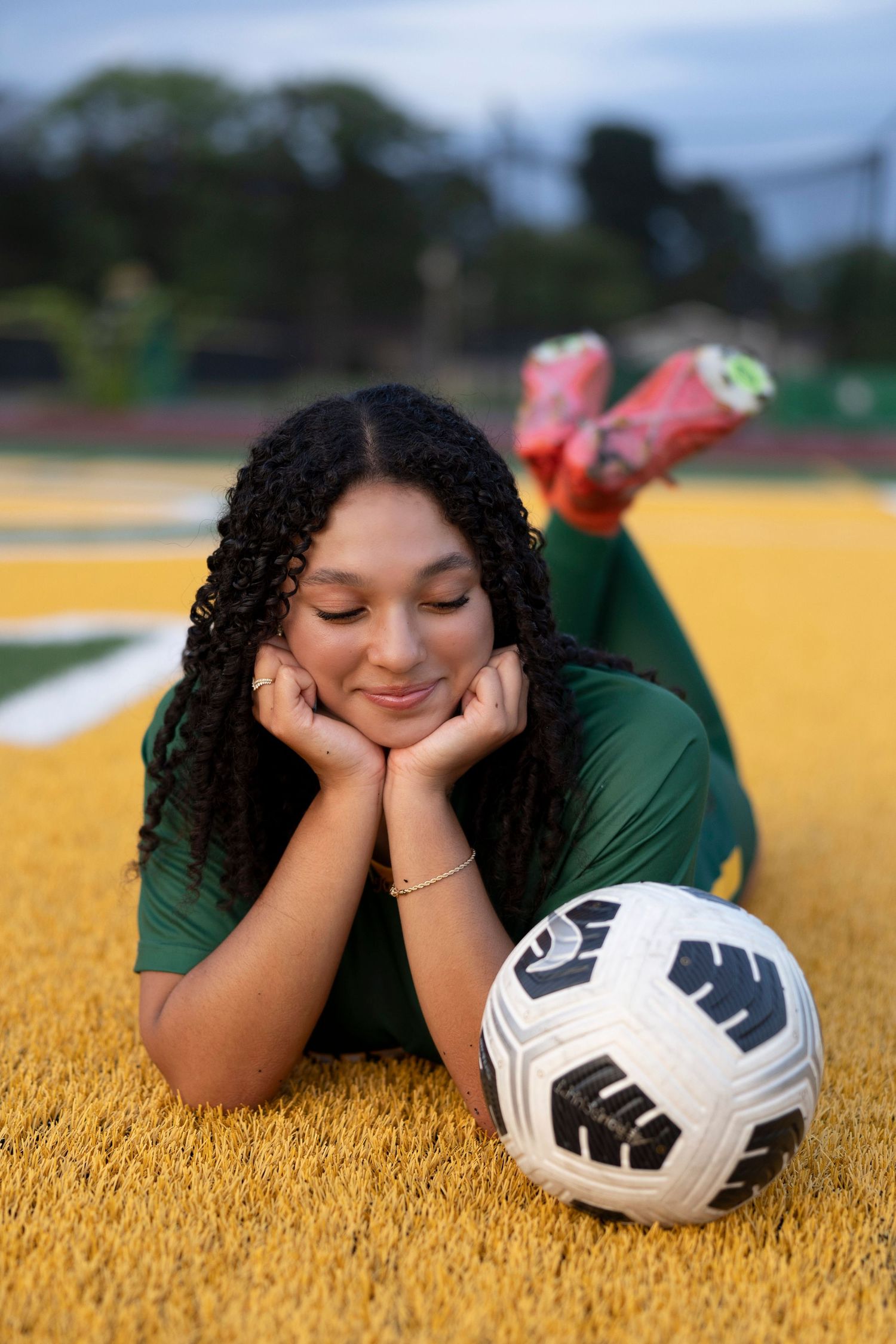 High school senior soccer player in green uniform lies on yellow turf field with soccer ball at Olympia High School.