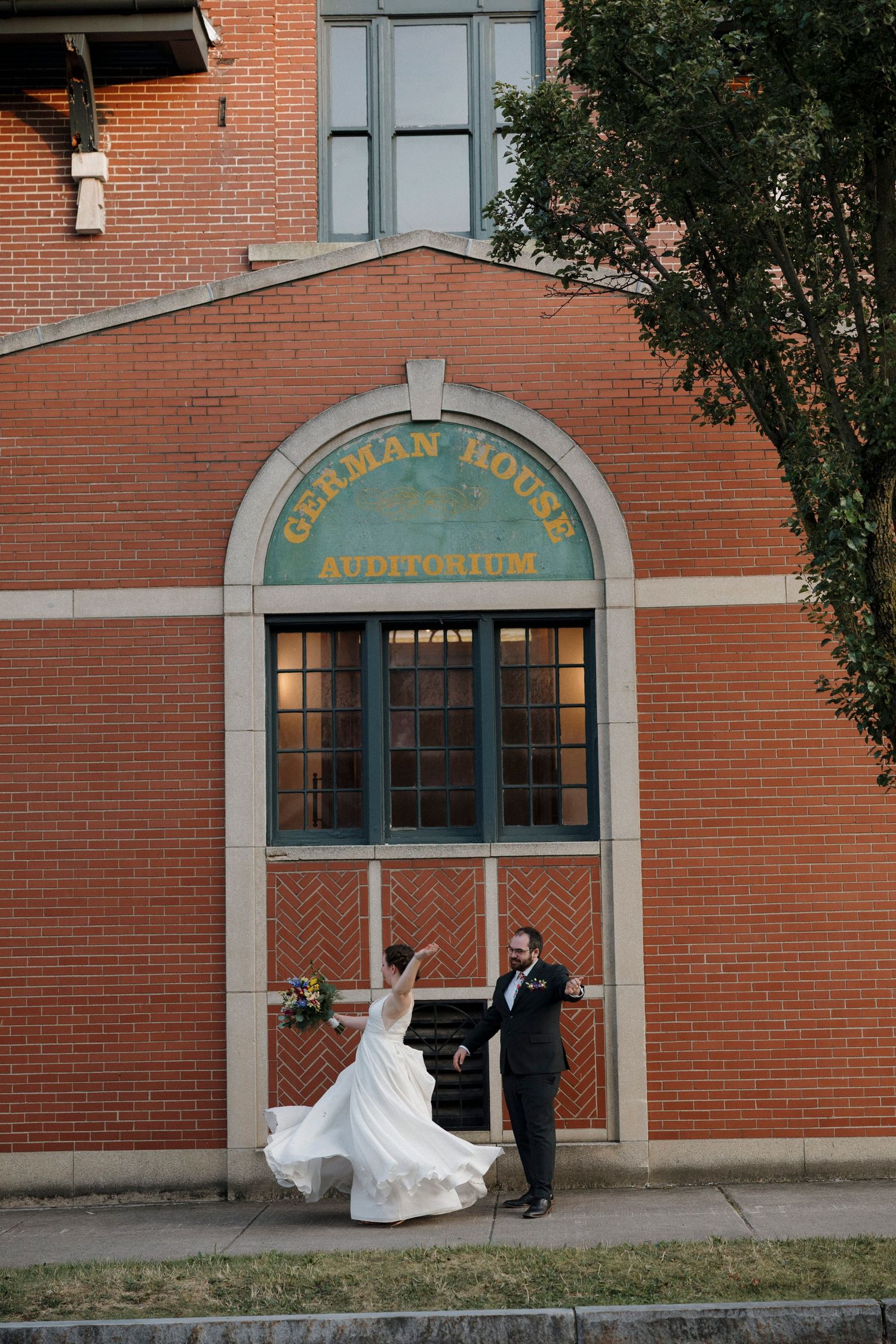 A couple twirls outside a brick auditorium building entrance during their wedding at the German house for wedding in Rochester, NY.