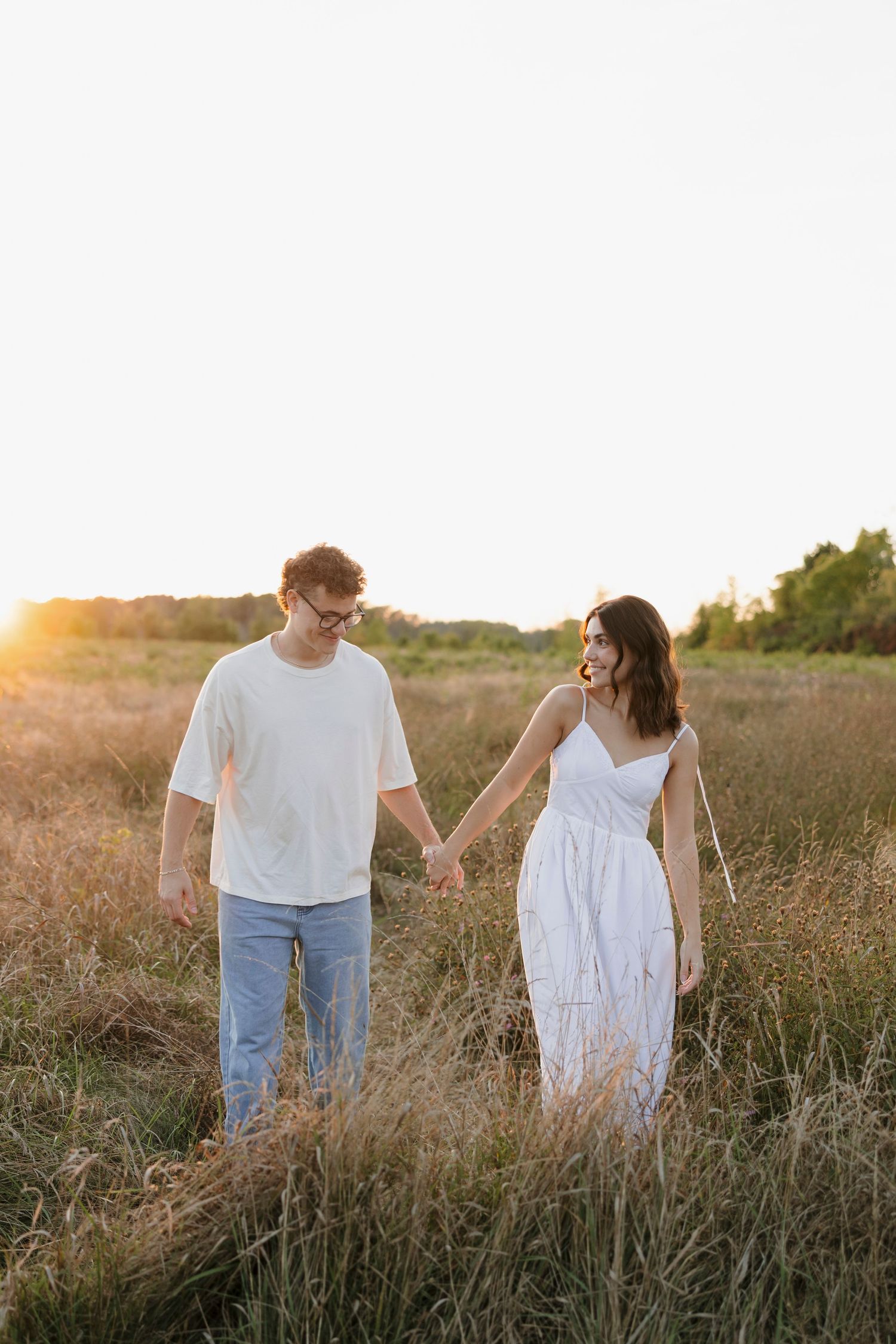 Couple in white outfits walking through golden sunset field in Hilton NY for engagement photos.