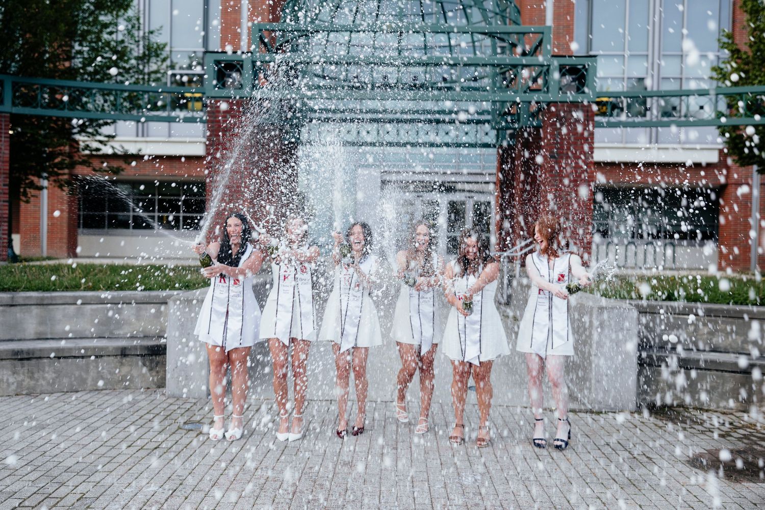 SUNY Geneseo graduation group celebrating with champagne sprays in front of a grand building entrance during graduation photos, Rochester NY
