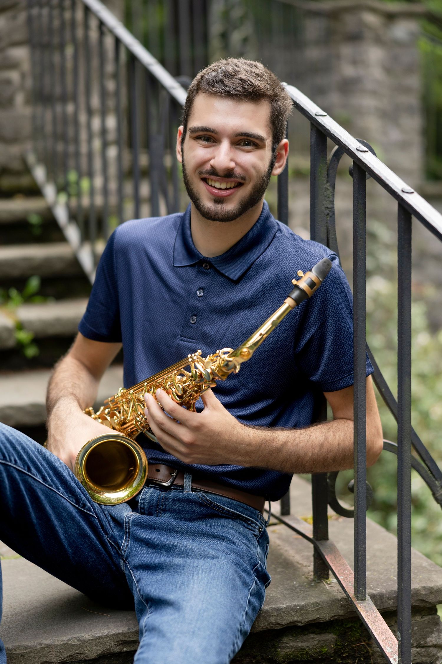High School senior boy sits on concrete steps playing saxophone in a navy blue polo and jeans.