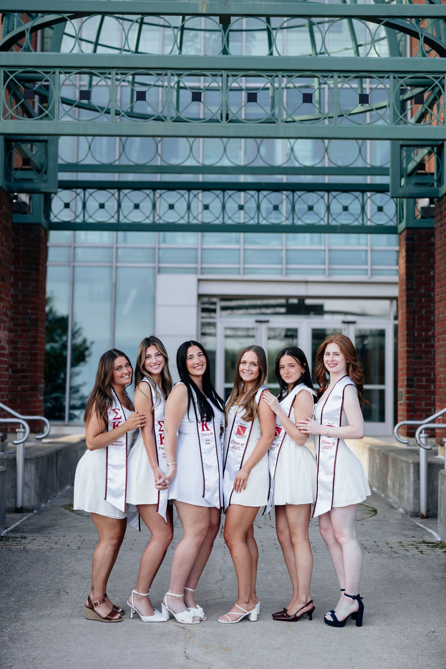 Group graduation portrait session in white dresses under a decorative glass and metal archway wearing white stolls.