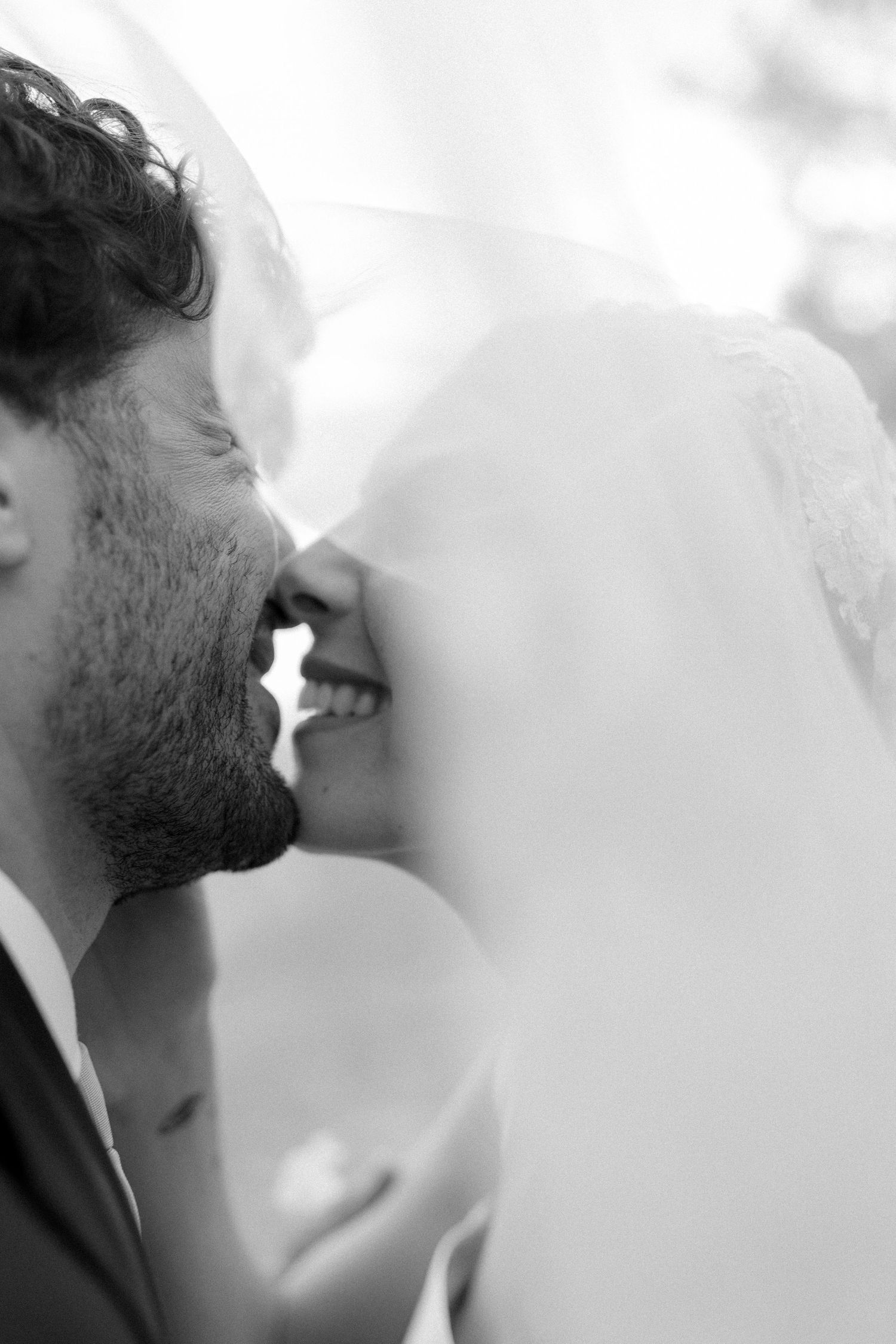 Black and white photo of a bride and groom sharing a romantic moment under a flowing veil on their wedding day.