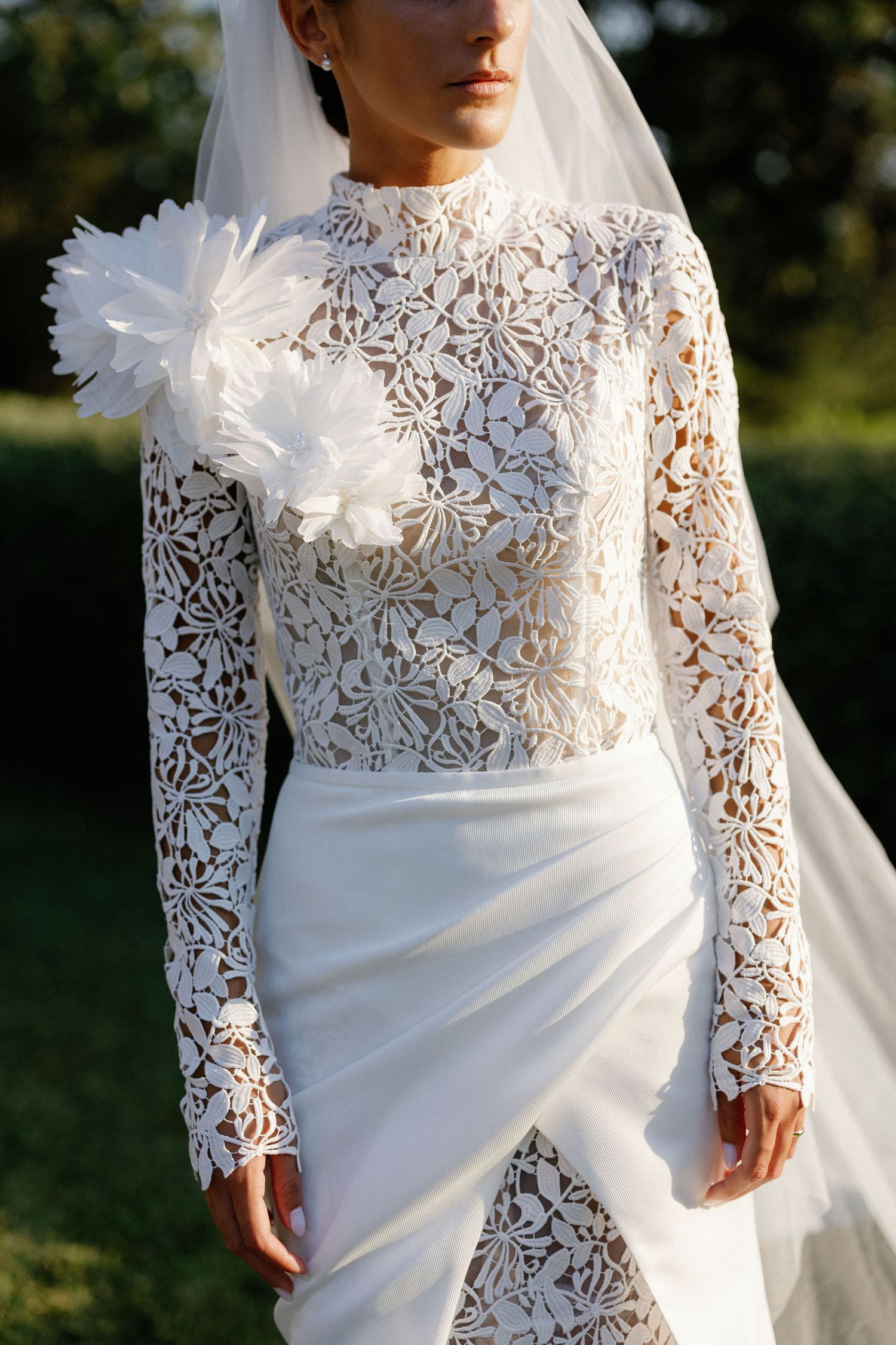 Bride in elegant white lace long-sleeve wedding gown with floral details and veil, standing outdoors.