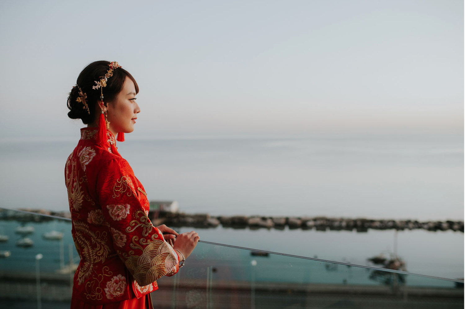 Woman in traditional red Chinese wedding qipao dress standing on balcony overlooking serene ocean at dusk.
