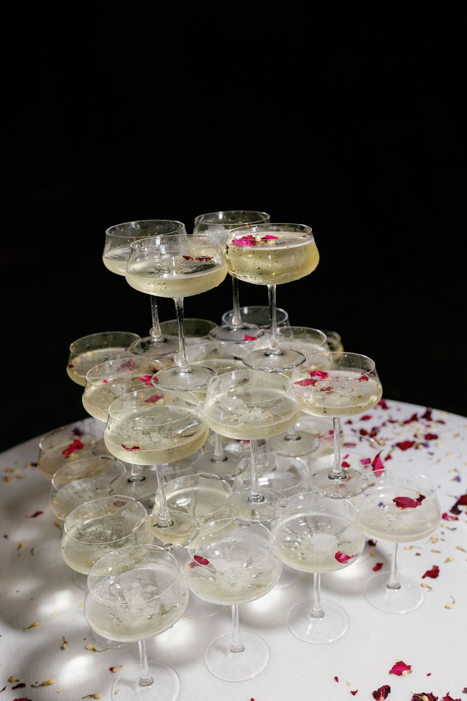 Elegant champagne tower with coupe glasses filled with sparkling wine, decorated with rose petals on a white table.