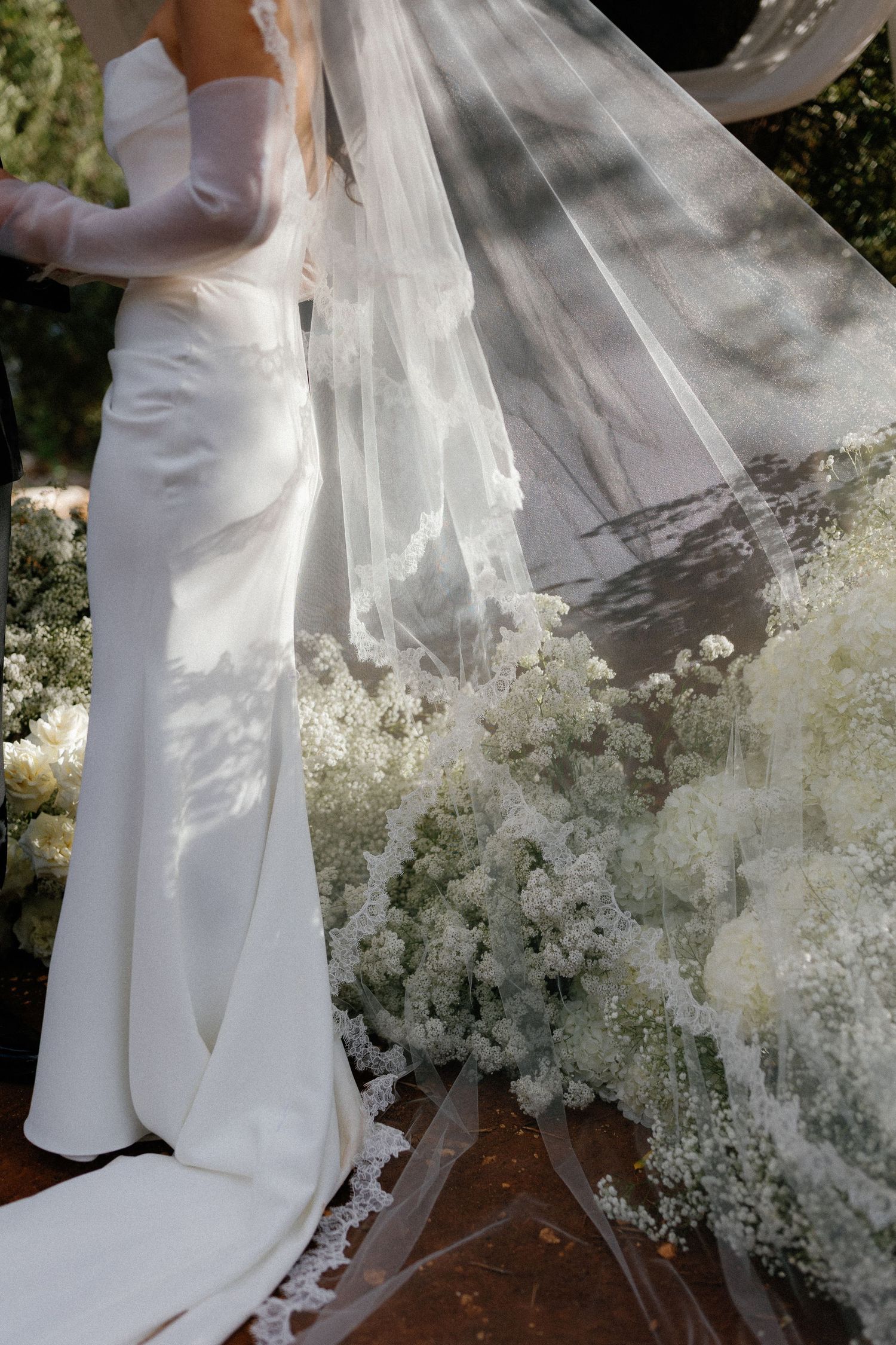 Bride in elegant white satin gown with long lace-trimmed veil surrounded by white baby's breath floral arrangements at outdoor wedding.