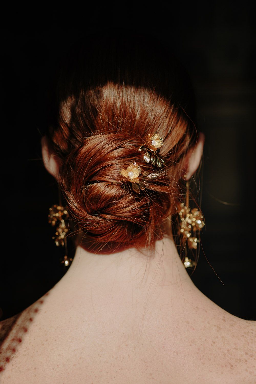 Elegant red hair updo with gold floral hair pins and matching chandelier earrings against dark background.