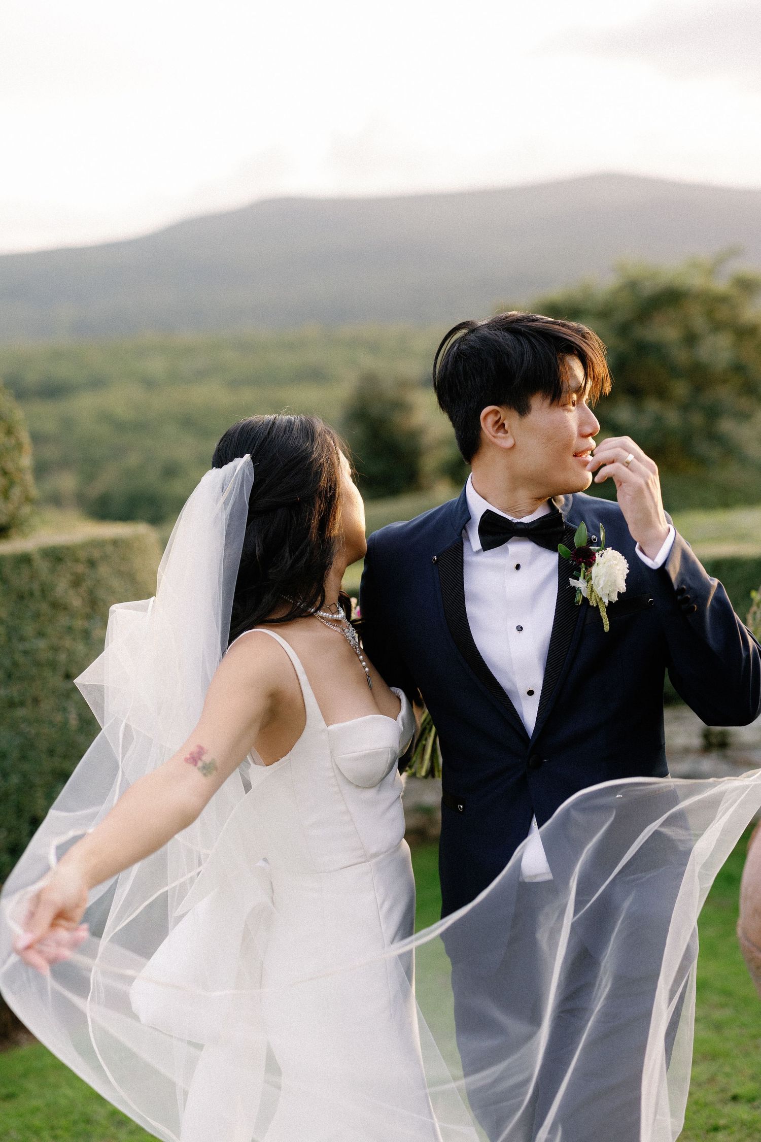 Asian bride and groom in tuxedo pose outdoors with veil flowing in wind, lush green hills in background.