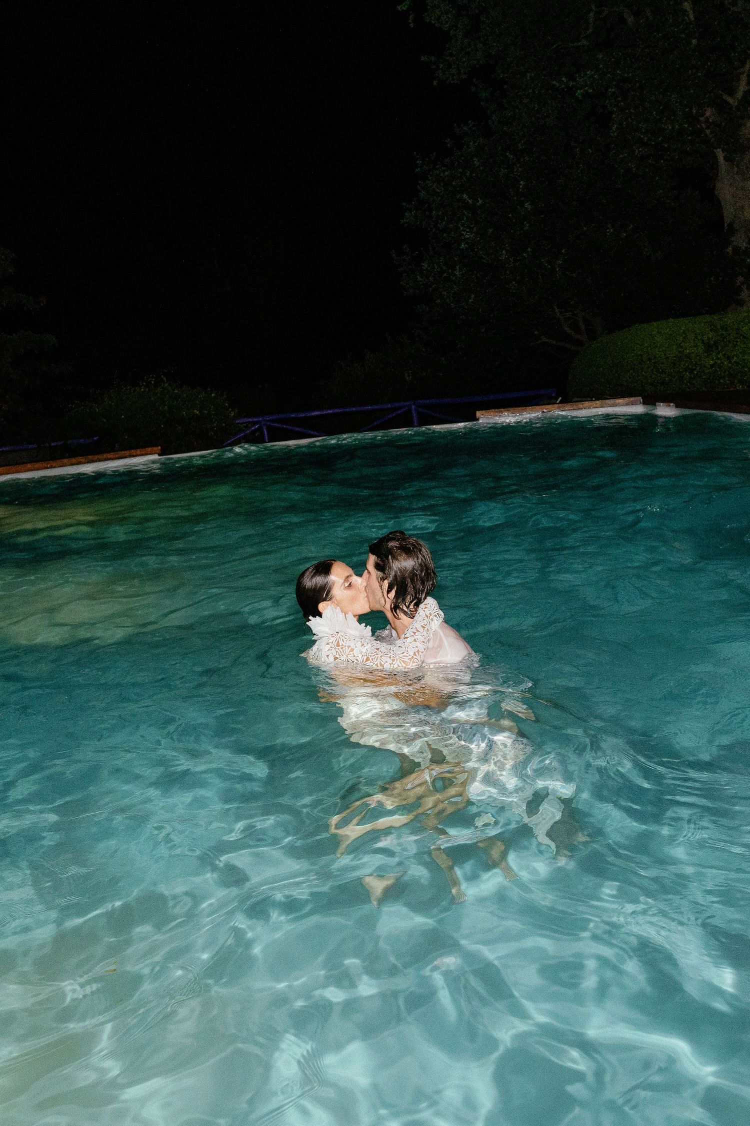 Two people kissing romantically while swimming in a turquoise pool at night.