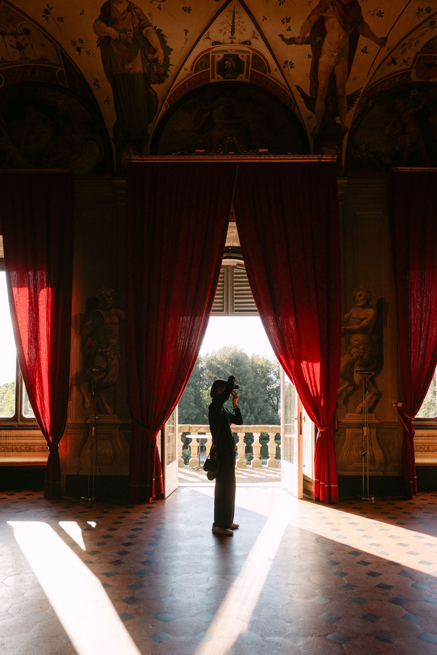 Silhouette of person photographing in ornate Italian villa with red curtains, frescoed ceiling, and sunlit marble floors.