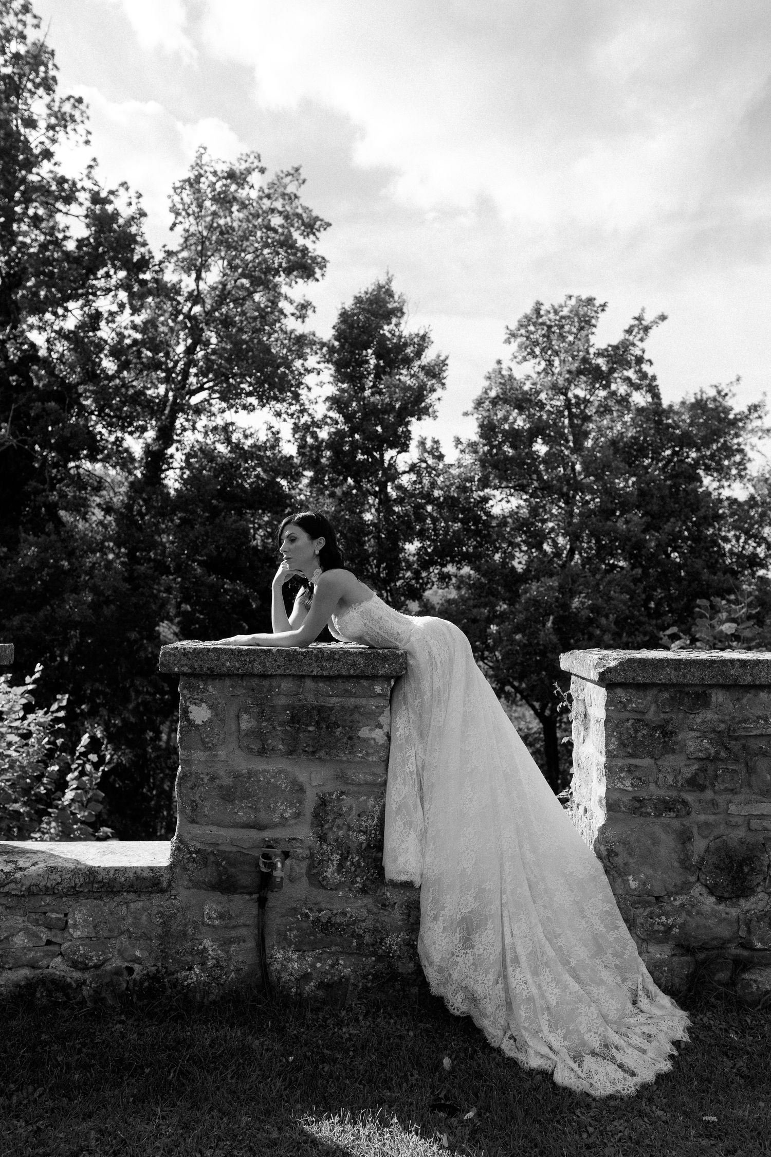 Bride in lace wedding gown leaning on stone wall surrounded by trees in elegant black and white outdoor portrait.