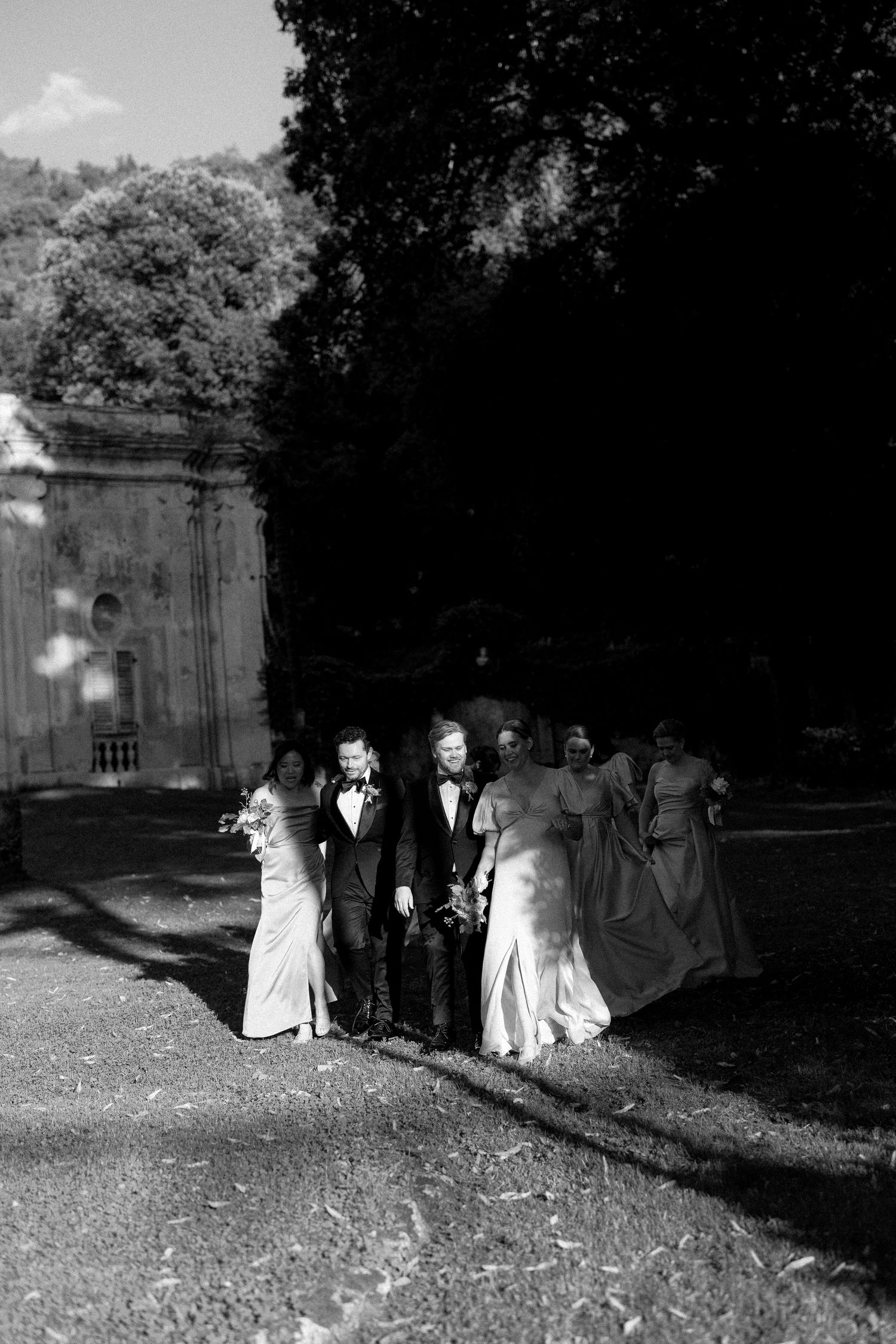 Black and white photo of wedding party walking outdoors near historic building, bride in white gown with groom and bridesmaids.