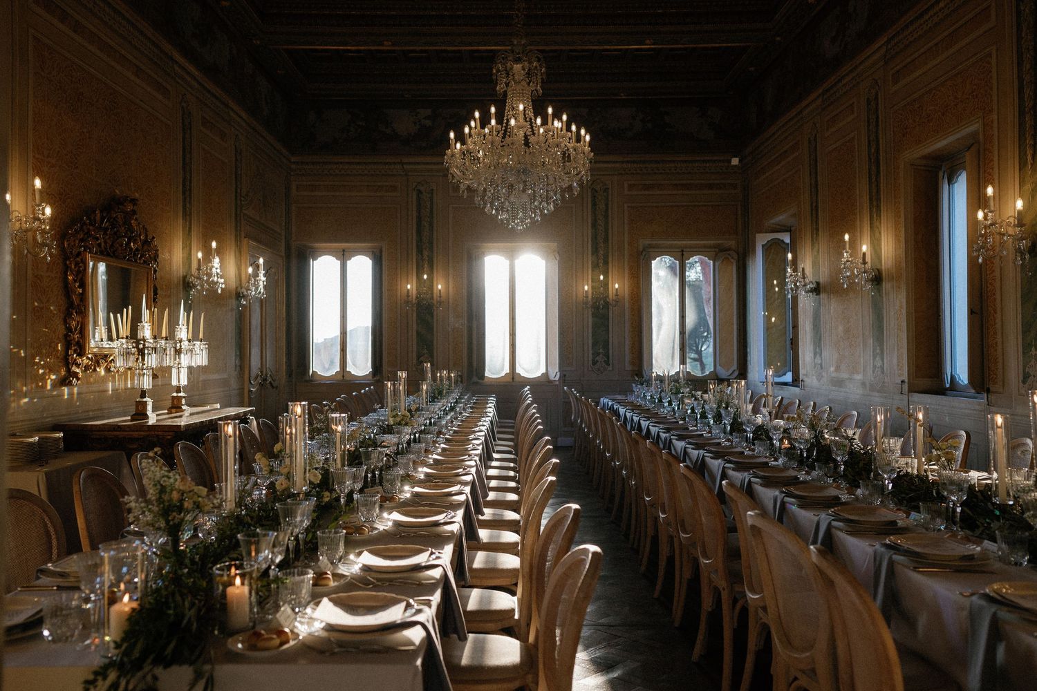 Elegant banquet hall with long dining tables, candles, floral centerpieces, and a grand chandelier in a historic venue.