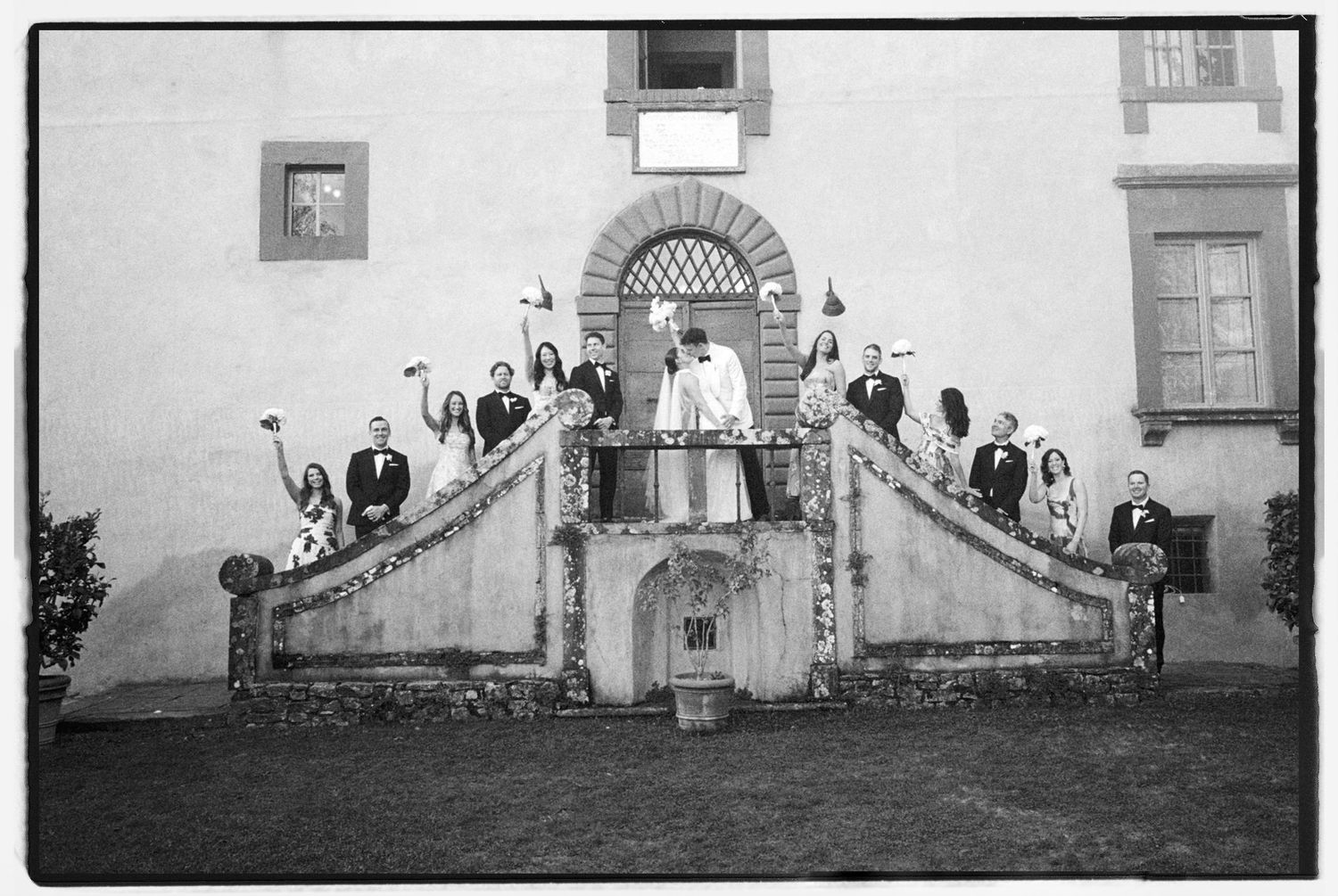 Black and white wedding photo of bridal party posed on ornate stone staircase balcony of historic villa.