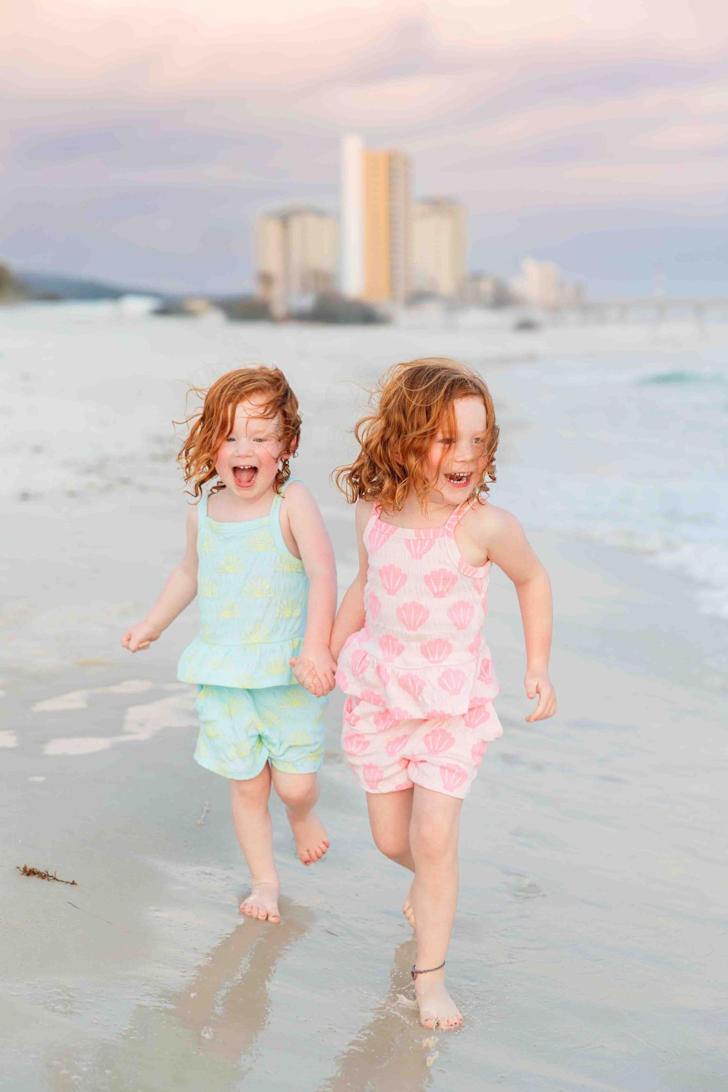Two children in pastel outfits running joyfully along a beach with high-rise buildings in the background.
