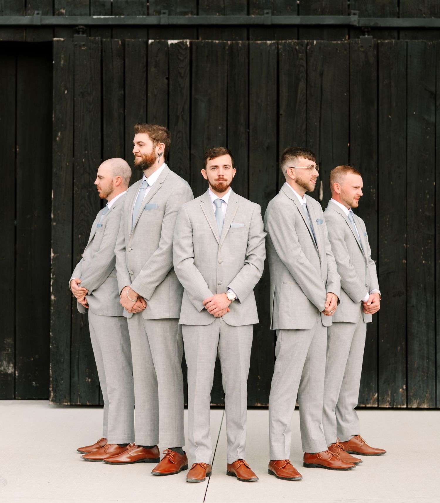 Wedding party in light gray suits lined up against a dark wooden wall.