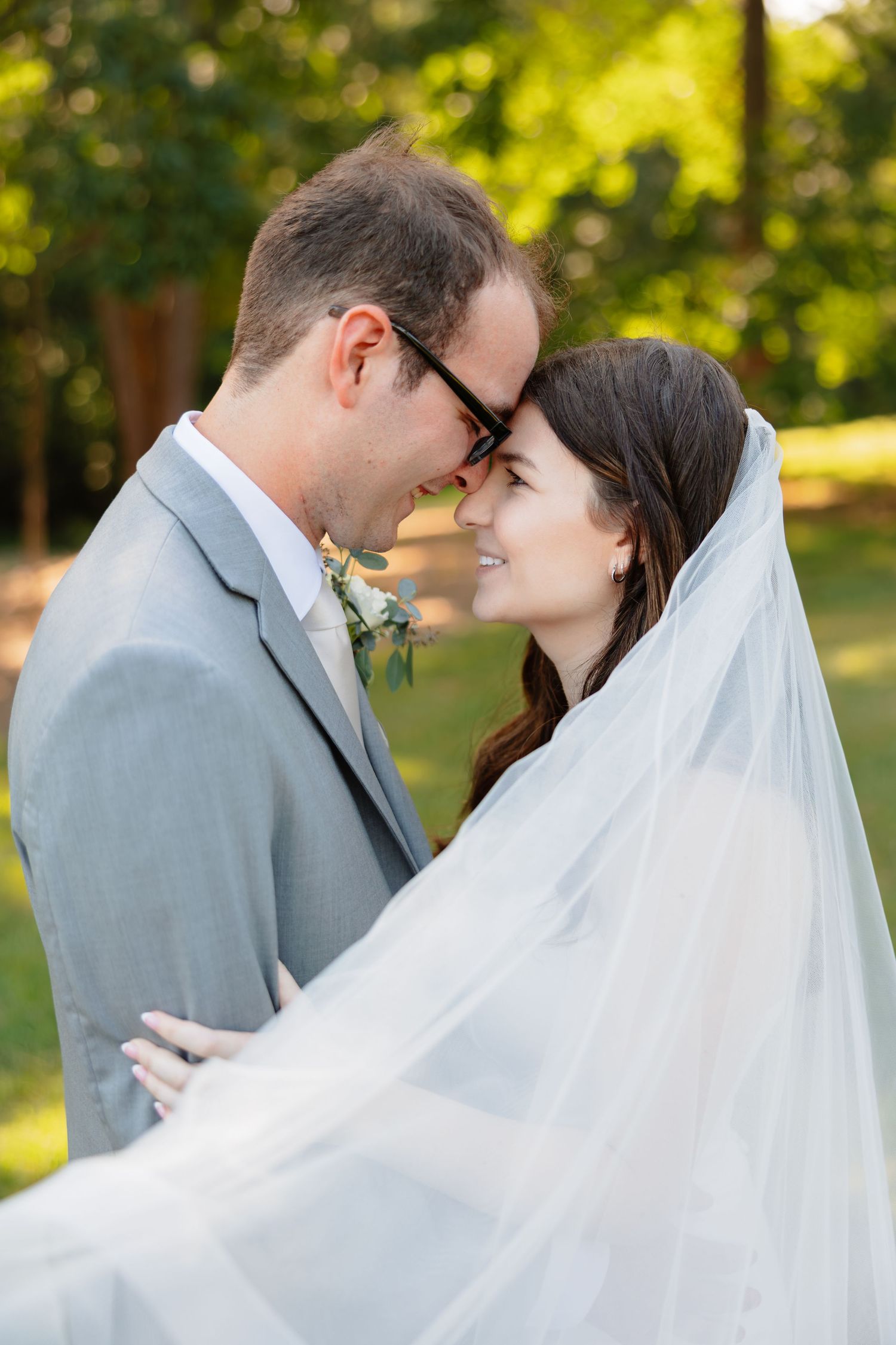 A bride and groom share a romantic forehead touch outdoors, with her white veil flowing around them in soft natural light.