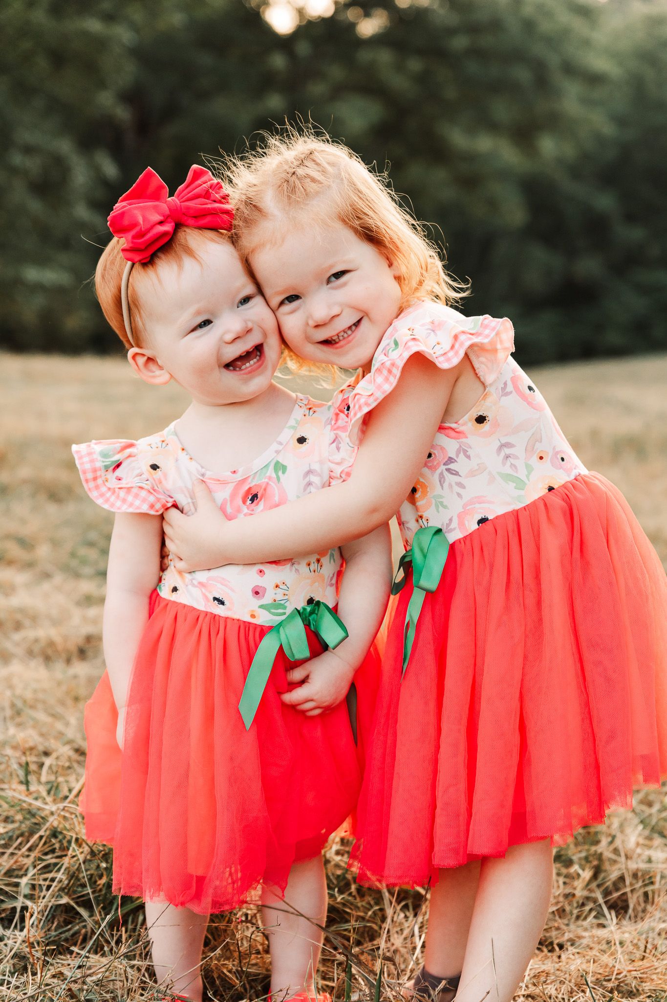 Two young children in matching coral-colored tulle dresses and bows share a sweet moment in a sunlit field.