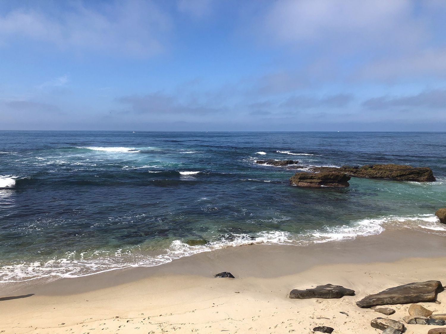 Panoramic view of a rocky beach coastline with crashing waves and blue ocean waters.