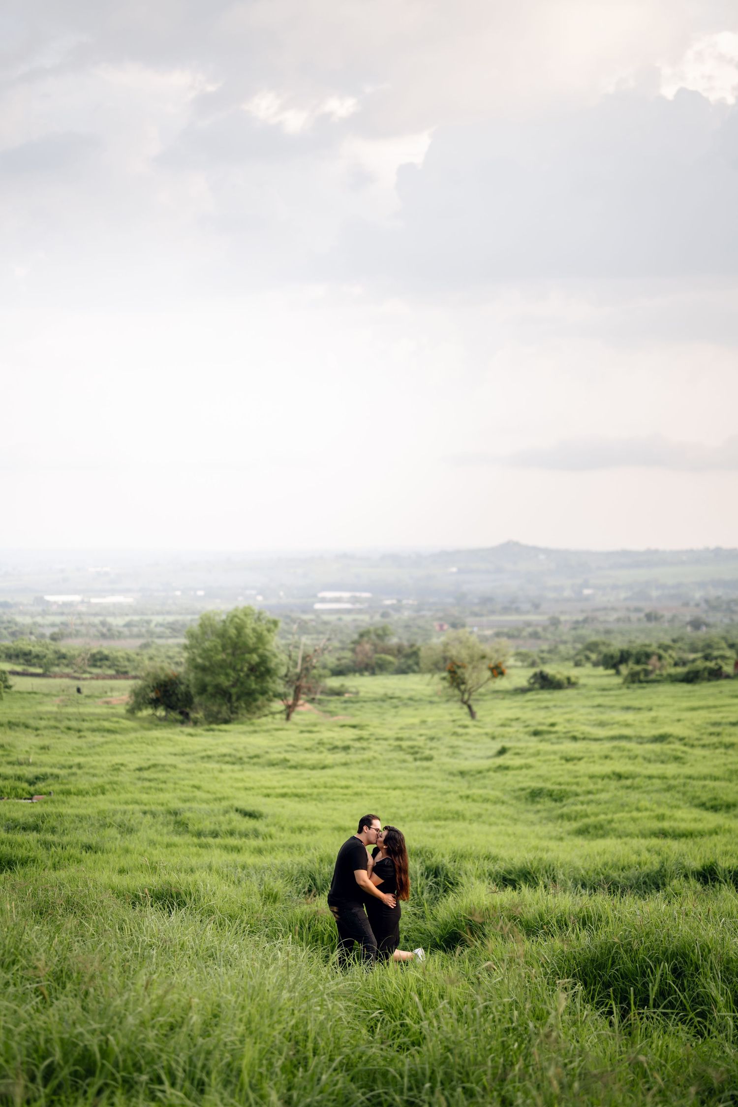 A couple shares a romantic moment in a lush green field with mountains in the background.